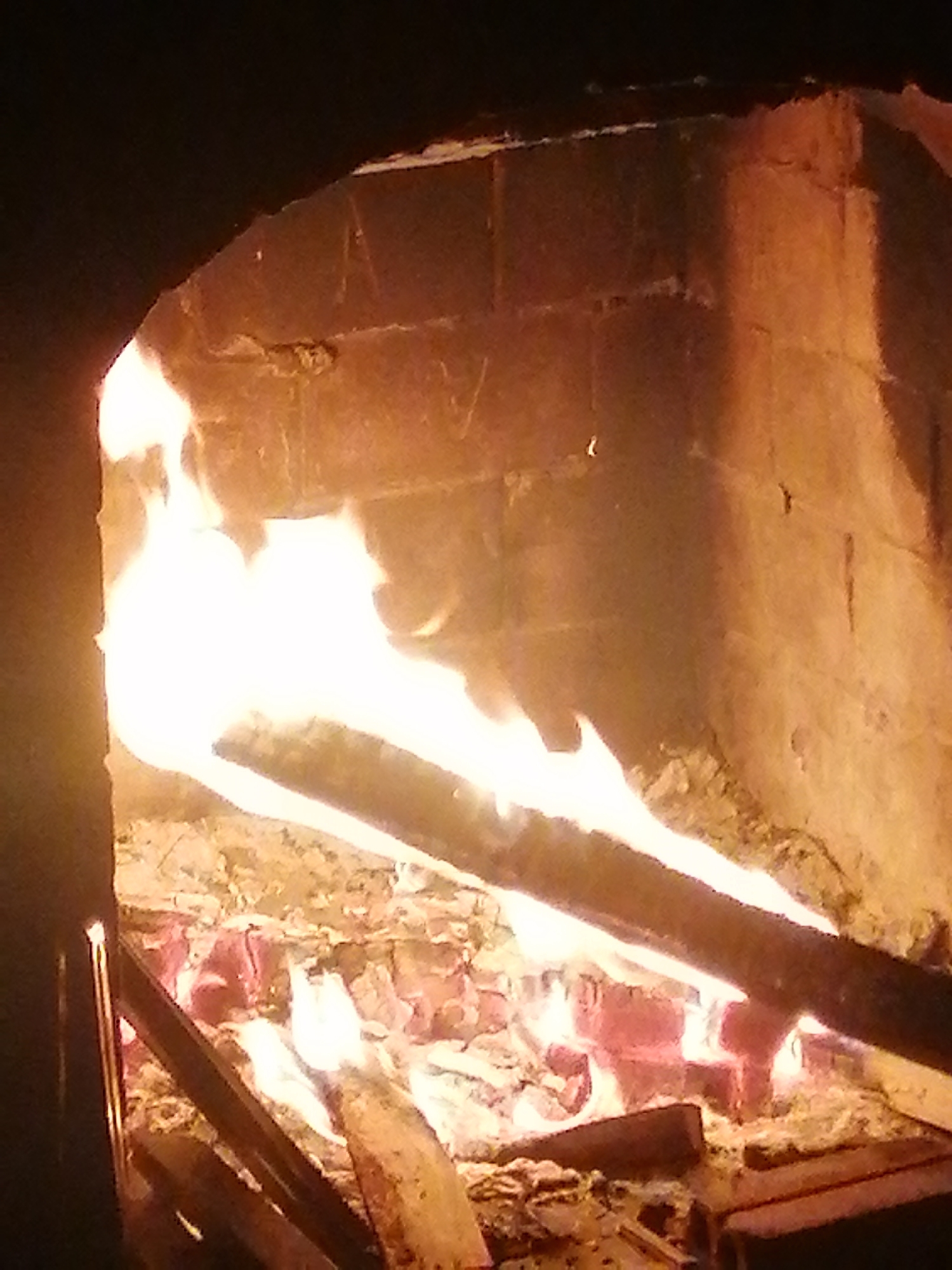 A warm-toned, close-up photograph captures a roaring fire within a fireplace. The scene is dominated by bright, energetic flames consuming several logs, casting a vibrant glow across the surrounding surfaces.
The fireplace is constructed from rough-hewn, light-colored stone or brick, which is strongly illuminated by the fire's light, revealing textures and shadows on its interior walls. A prominent, elongated log stretches horizontally across the middle of the frame, actively engulfed in bright white and yellow flames that extend upwards and around it. Below this, a bed of glowing red and orange embers, interspersed with ash and smaller, partially burned wood pieces, forms the base of the fire. A dark, possibly metallic, grate or fire iron is visible in the lower-left corner of the hearth.
The activity taking place is a natural wood fire burning intensely. The fire serves as the primary light source in the image, suggesting an indoor setting, likely during the evening or night. The overall impression is one of warmth, comfort, and a cozy atmosphere. There are no people visible in the frame, and no discernible text is present. While the location context is Lạc Dương, Vietnam, the visual details within the image itself do not offer specific architectural or environmental cues unique to that region.