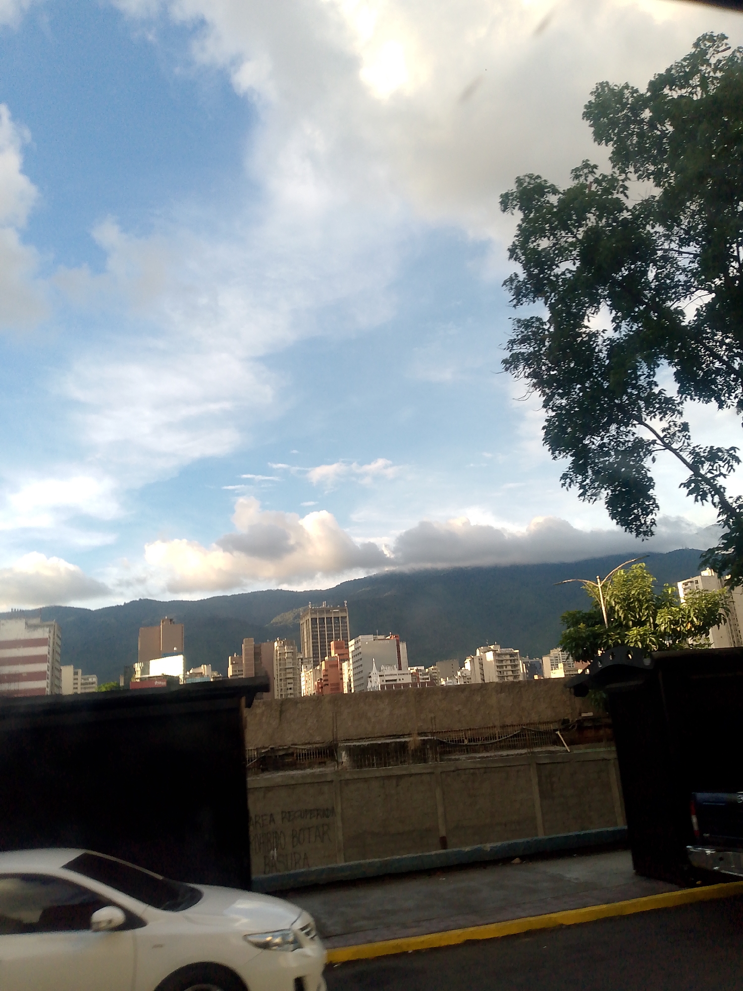The image shows a street scene in Caracas, Venezuela, under a cloudy sky with a mountain range in the background. Buildings of varying heights rise from the cityscape. In the foreground, a white car is partially visible, with a grey concrete wall behind it, on which there is some graffiti. A black box-like structure partially obscures the view, and a tree with dark green foliage is on the right side. The street has a yellow curb.