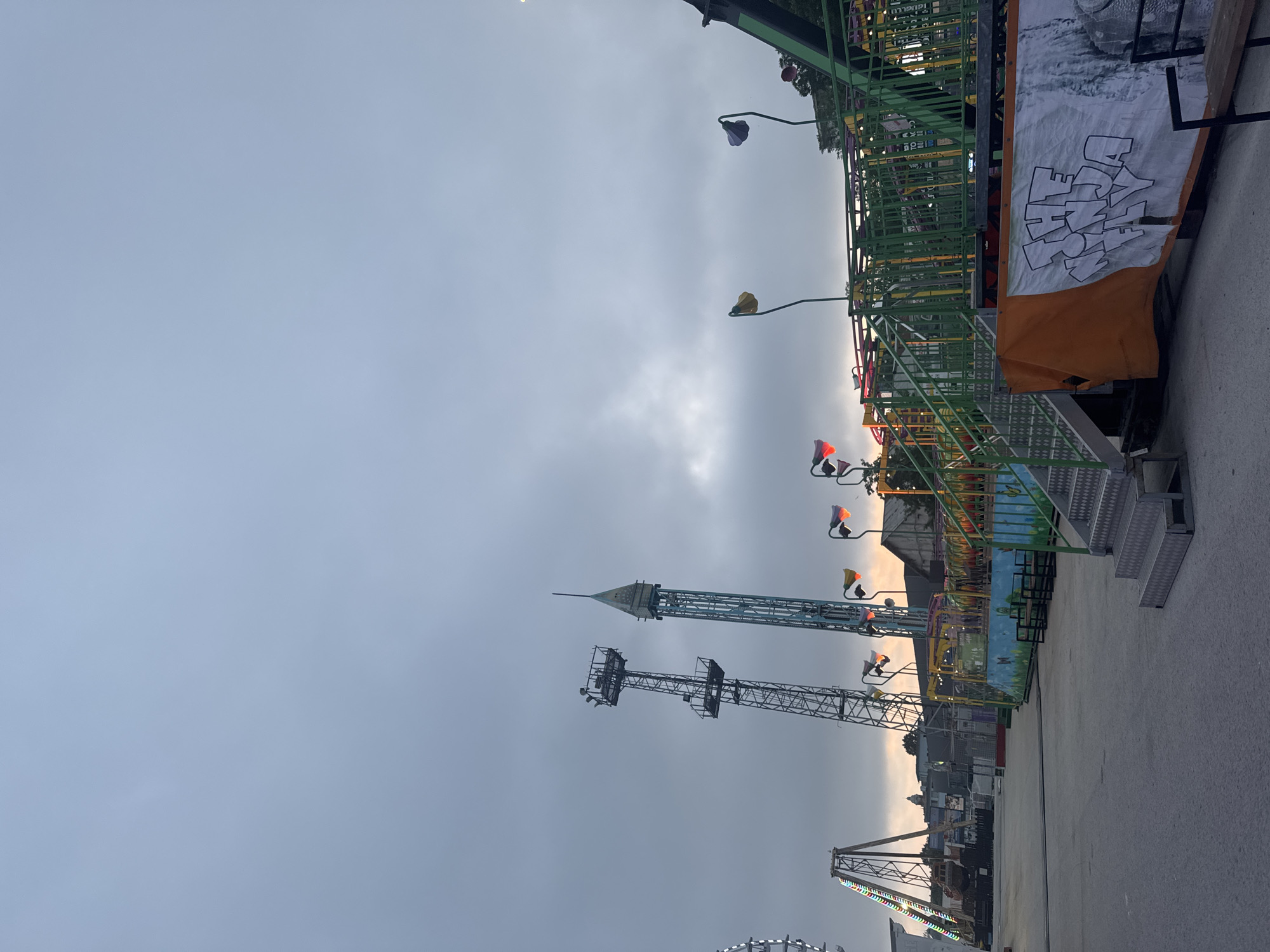 The image depicts an amusement park located in Varna, Bulgaria, at dusk under an overcast, gray sky. The foreground features an empty asphalt ground. To the right, a metal staircase with diamond plate steps ascends to a large amusement ride structure, primarily characterized by green railings and yellow accents. Below this ride, a banner is affixed, displaying an orange section on the left and a white section on the right with black graffiti-style text that reads "THE NINJA FLY." Below the banner, partially visible benches are present, and the ride's base is adorned with blue and light green panels featuring faint butterfly and flower motifs.
In the midground, multiple amusement park attractions are visible. Two prominent tall towers rise into the sky: one on the left is a dark, lattice-structured metal tower with multiple platforms, while the other to its right is a slender, light blue/teal tower with a pointed top. Decorative lights resembling blooming flowers, in red, yellow, and purple hues, illuminate parts of the immediate ride areas. To the far left, a section of a large Ferris wheel with bright white lights is visible, alongside another swinging or pendulum-type ride structure.
The background reveals a distant urban landscape, likely parts of Varna, with buildings silhouetted against a slightly lighter horizon where the sun appears to be setting behind the clouds. Trees and other foliage are visible on the far right, bordering the amusement park. No individuals are discernible within the frame. Artificial lights from the rides and decorative elements are active, contrasting with the diminishing natural light of the evening sky.