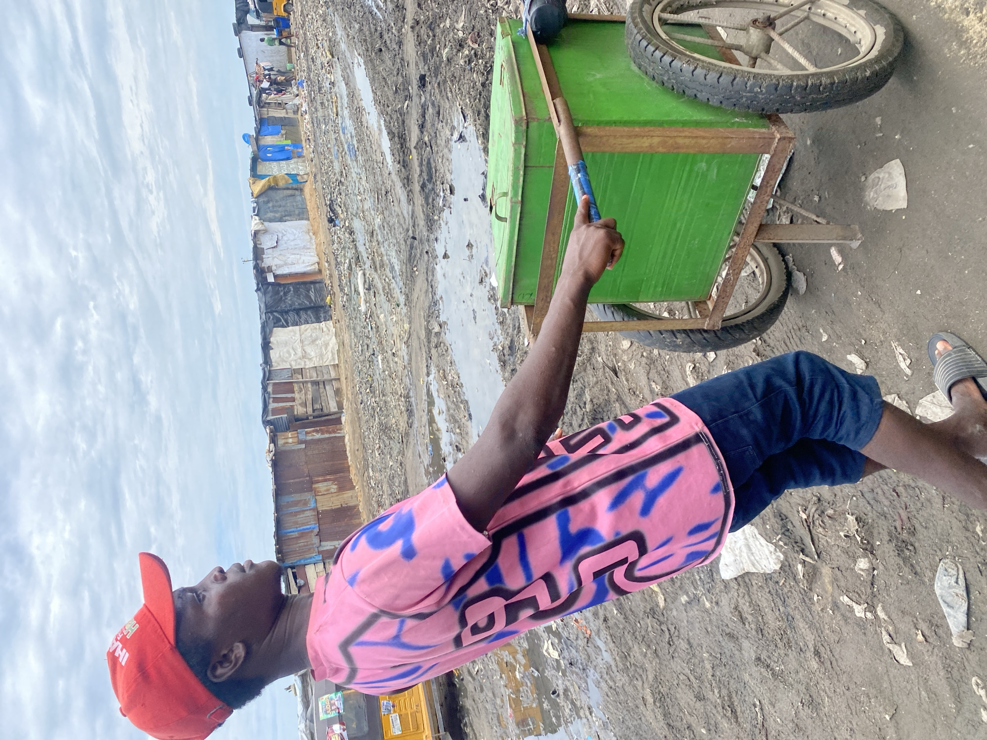 In the city of Ado, Nigeria, a young boy wearing a red baseball cap and a pink t-shirt with blue and black graffiti-style markings pushes a green, metal cart on two wheels. The cart appears to be a cooler or storage box. He is dressed in dark blue shorts. The environment is a slum with makeshift structures made of corrugated metal and other salvaged materials. The ground is muddy and littered with debris, indicating a poorly maintained area. In the background, other structures and a yellow tricycle are visible, as well as a person in a green shirt. The sky is overcast.