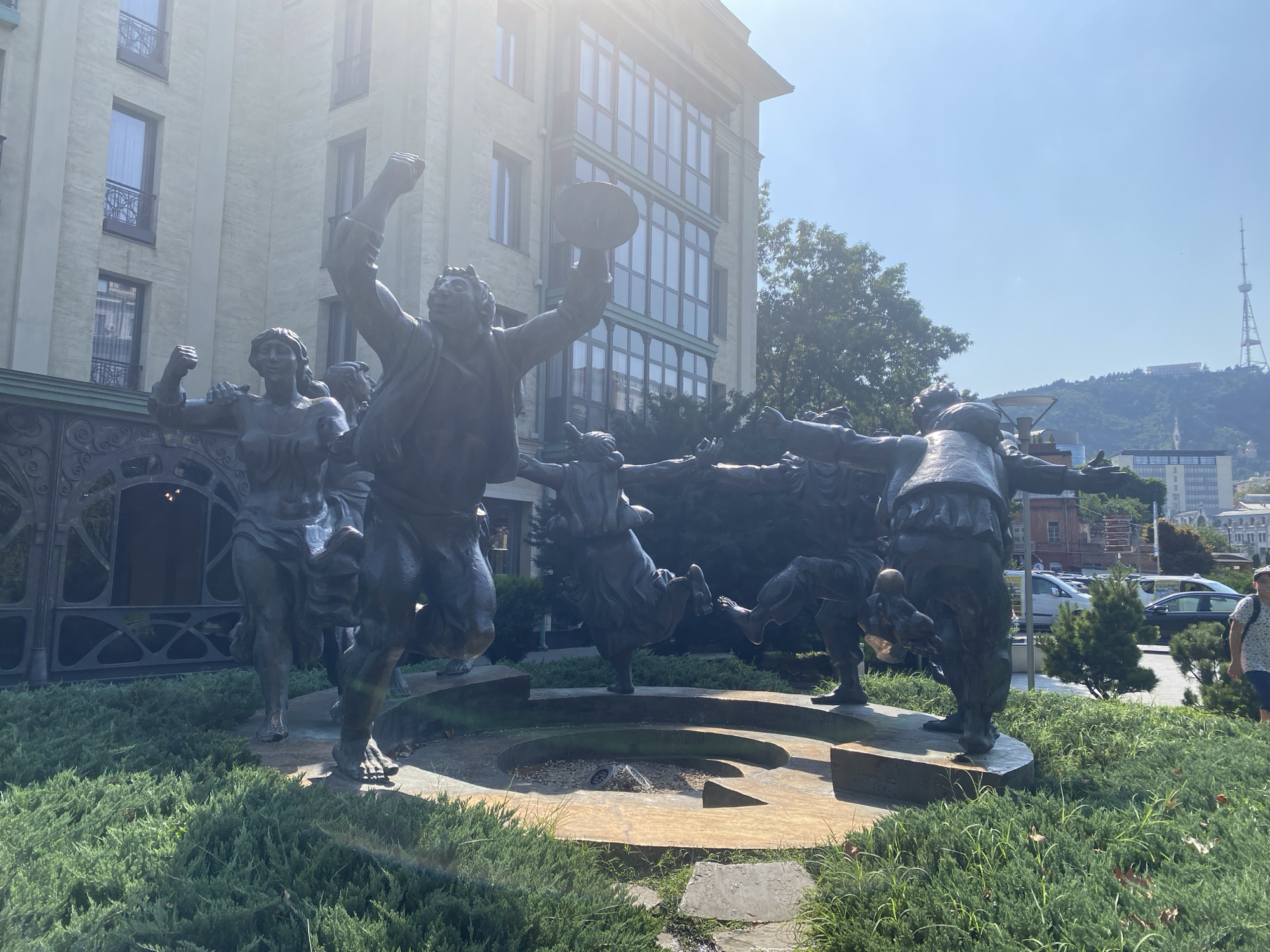 The image captures a bronze sculpture depicting several figures in dynamic, dancing poses, located in Tbilisi, Georgia. The sculpture is situated in front of a light-colored building with a modern facade featuring large windows. In the background, a television tower is visible on a hill, characteristic of the Tbilisi skyline. The scene is illuminated by bright sunlight, creating lens flare in the upper left portion of the image. The foreground consists of lush green bushes and some stone pathways. A few people are visible in the background, including one person walking towards the right side of the frame. The overall environment suggests a public outdoor space, possibly a plaza or park area.
