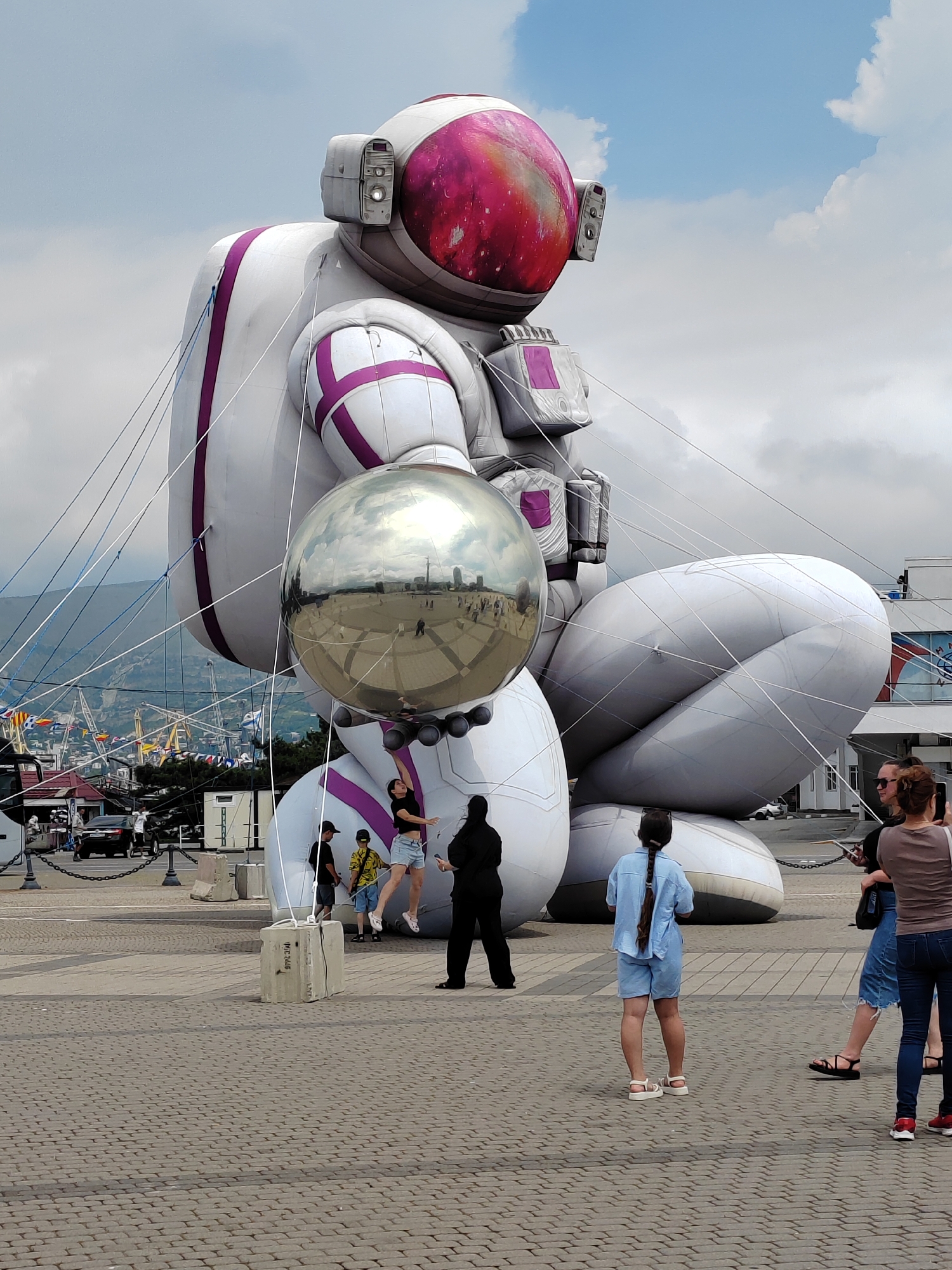 An inflatable astronaut figure, approximately 10-15 meters tall, is displayed outdoors on a paved area. The astronaut is white with purple accents and holds a large, reflective sphere in its left hand. Several people, including adults and children, are present in the foreground and midground, observing the sculpture. Some individuals are taking photos. The background reveals a city skyline with cranes and buildings, suggesting an urban or port area. The sky is overcast with some visible clouds.