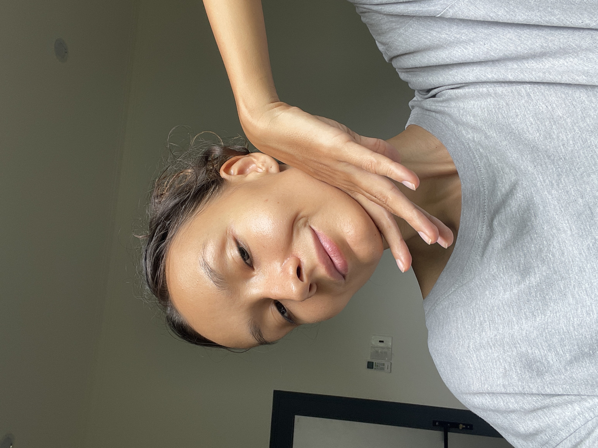A serene portrait captures a woman with a gentle, slightly upward gaze and a soft smile, her head tilted to one side. She has dark hair, a medium complexion, and full lips. Her right hand, with neatly manicured nails, is gracefully positioned against her cheek and neck, with her arm bent and partially visible. She is wearing a light gray, textured t-shirt, which shows soft folds across her shoulder and chest area.
The background consists of a plain, muted grayish-green wall, suggesting an indoor setting. In the lower portion of the image, a dark rectangular frame, possibly a mirror or picture, is visible. Above it on the wall, there are some indistinct rectangular objects, which could be a light switch or thermostat panel. The lighting is soft and even, illuminating her face and skin with a natural glow. The overall impression is one of calm and contemplation. The scene is situated in Mũi Né, Vietnam. No visible text is present.