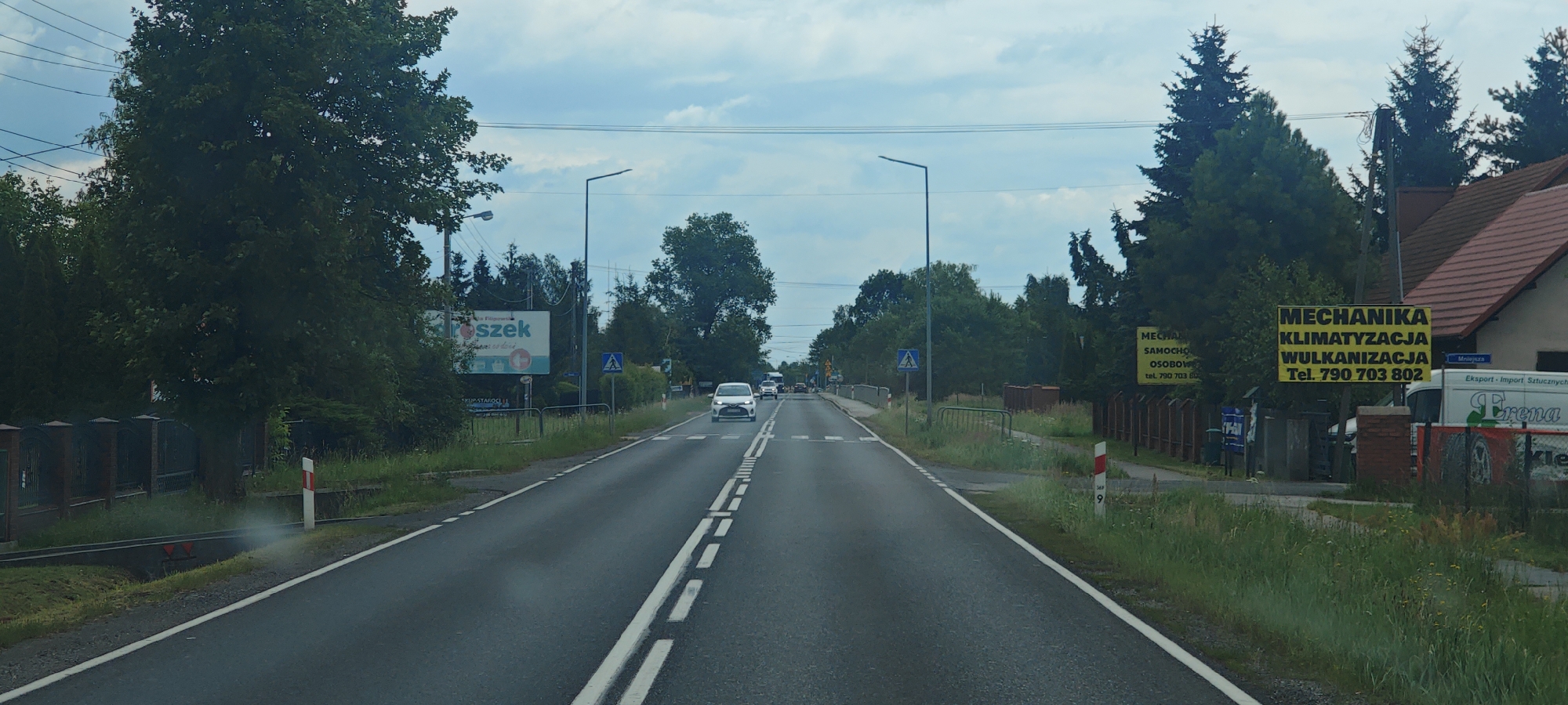 The image shows a straight, paved road with white dashed lines dividing the lanes. The road is bordered by grassy areas and trees on both sides. There are streetlights and power lines visible overhead.
On the left side of the road, there is a billboard that appears to advertise a business. Further down the road on the left, there is a black metal fence.
On the right side of the road, there are several yellow billboards advertising "MECHANIKA KLIMATYZACJA WULKANIZACJA" and a phone number. A white van with "Crena" written on it is parked near these billboards, and a portion of what appears to be a tire is visible. There is also a residential building with a tiled roof.
Several cars are visible on the road, moving away from the camera. One white car is in the foreground. There are also road signs, including a pedestrian crossing sign. The sky is cloudy.
The scene depicts a road in a semi-rural or suburban area, likely in Wola Filipowska, Poland, given the Polish text on the billboards and the overall environment. There are no people clearly visible in the image. The overall impression is of a typical roadside scene with commercial advertisements and passing traffic.
