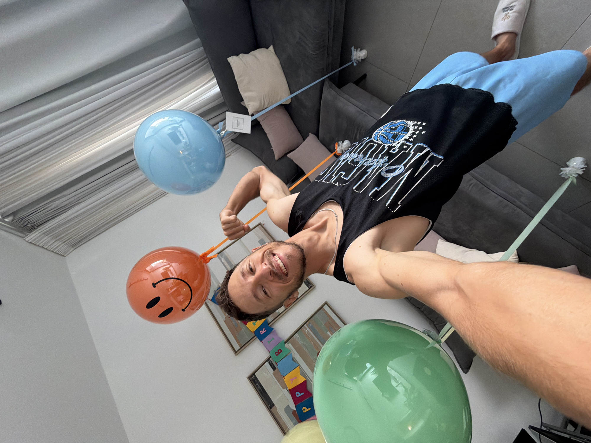 A man is taking a selfie in a room with decorations for a birthday celebration. He is holding the strings of two balloons, one orange with a smiley face and another light blue. To his left, a large green balloon is visible. Behind him, a banner that spells out "BIRTHDAY" is hanging, along with two framed pictures on the wall. The man is wearing a black tank top and light blue shorts, and he is smiling at the camera. The room appears to be a living area, with a large grey sofa and a curtain in the background. Based on the user's prompt, this scene is located in Tel-Aviv, Israel.