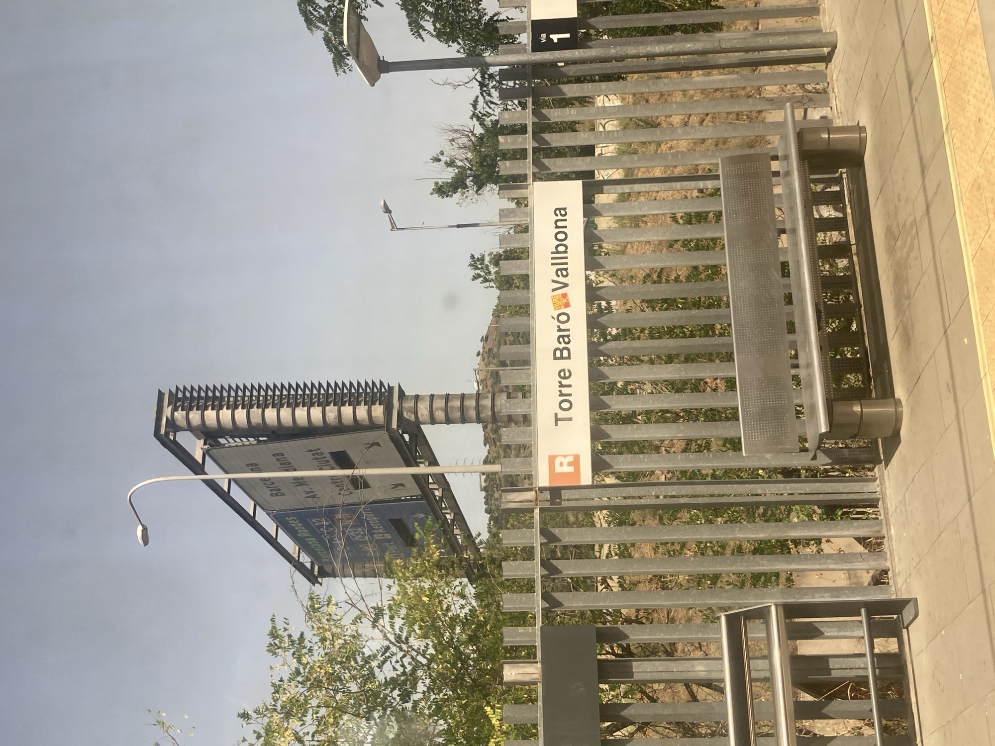 The image captures a section of the Montcada i Reixac train station in Spain. In the foreground, a metallic fence runs horizontally across the frame, separating the platform from the tracks. A modern, metallic bench is situated on the platform. Above the fence, a station sign reads "R Torre Baró Vallbona" with a small orange and yellow emblem to the left of "Vallbona". To the right of the sign, a black square indicates "via 1". In the background, a large billboard with directional information for various destinations, including "Barcelona" and "Aeroport", is visible. Green foliage and trees are present behind the fence and billboard. The sky is clear and light blue.
