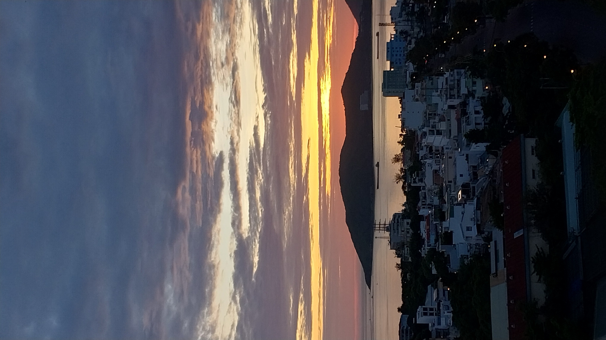 A vertical, high-angle shot captures a scenic view of a coastal city at dusk. The sky is a dramatic blend of deep blues, purples, and oranges, with streaks of yellow and pink from the setting sun painting the horizon. Wispy clouds are scattered across the sky, adding texture to the twilight atmosphere.
In the foreground, the dark silhouettes of trees frame the lower portion of the image. Below the trees, a dense urban landscape unfolds, characterized by a collection of buildings with white and light-colored facades. These structures range from multi-story apartment buildings to smaller houses, many with flat roofs. A paved road with a series of glowing streetlights winds through the city, suggesting the transition into evening.
Dominating the midground is a calm body of water, likely a bay or ocean, reflecting the soft colors of the sky. On the water, a few dark shapes of ships or boats are visible. Beyond the water, a prominent, dark, mountainous island rises majestically, its peak softened by the fading light. The island appears uninhabited, its slopes covered in vegetation.
The overall impression is one of a tranquil coastal city bathed in the warm, fading light of sunset. The scene is devoid of any discernible people or specific landmarks that would pinpoint an exact location, but the architecture and the presence of the sea and mountains suggest a Southeast Asian coastal setting.