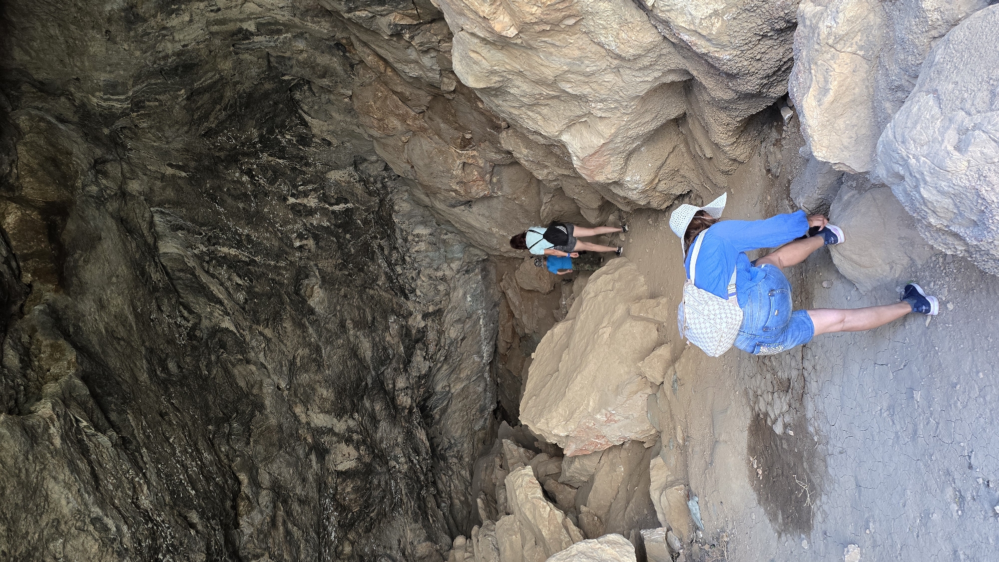 The image depicts the interior of a cave, with rough, textured rock formations dominating the scene. In the foreground, a person wearing denim shorts, a blue long-sleeved shirt, a white patterned backpack, and a white wide-brimmed hat is bent over, adjusting their dark sneakers on a rock. Further into the cave, two other individuals are visible. One, wearing a light blue shirt and dark shorts, stands facing away from the camera with a black backpack. Next to them, a child in a blue shirt is also facing away. The cave floor is composed of dirt and gravel, with several large boulders scattered throughout. The cave's ceiling is a mass of dark, uneven rock. The location is identified as Terekty, Kazakhstan.