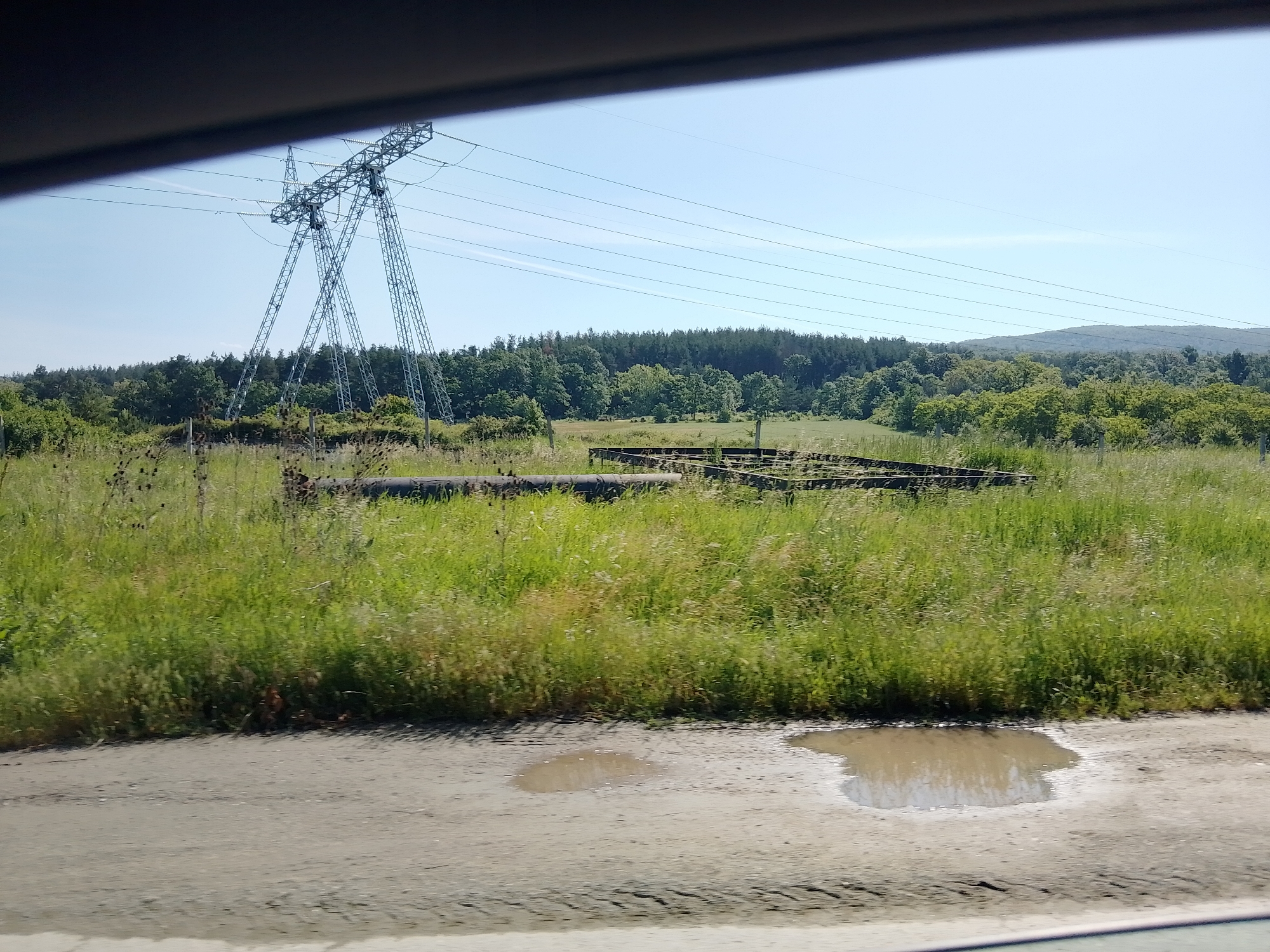 The image displays a rural landscape captured from what appears to be a moving vehicle, indicated by the dark, blurred frame at the top and bottom. In the foreground, a dirt road or shoulder is visible, featuring two prominent muddy puddles and faint tire tracks.
The midground is occupied by a sprawling field of tall, wild green grass, with some patches of drier, light brown vegetation. Dominating the left side of the field is a large, metallic lattice-work pylon, characteristic of a high-voltage electrical transmission tower, with numerous power lines extending horizontally across the sky. Further within the field, a dark, cylindrical object resembling a pipe lies on the ground, and to its right, a low, rectangular structure, possibly a metal or wooden frame, is partially obscured by the grass.
In the background, a dense forest composed of various trees forms a thick green line across the horizon. Beyond the forest on the right, a range of distant, rolling hills or mountains is faintly visible under a clear, bright blue sky. A single faint, wispy cloud streak can be seen in the sky.
The scene is brightly lit by natural sunlight, suggesting a daytime setting, likely late morning or afternoon. The weather appears clear and sunny. No people or specific activities are present, and there is no visible text in the image.