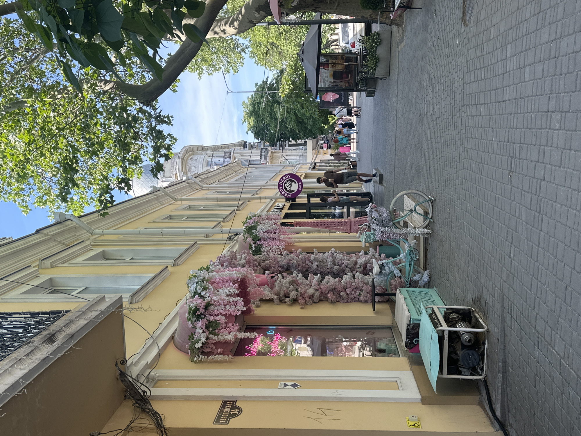 This image captures a daytime scene on a pedestrian street, identified by a sign as "ВУЛИЦЯ ЄВРОПЕЙСЬКА 14" (Yevropeiska Street 14), likely in an unknown Ukrainian city. The weather is clear and sunny, indicated by bright light and blue sky.
The left side of the street features a multi-story building with a pale yellow facade, adorned with white window frames and decorative architectural elements, including a balcony with a black wrought-iron railing on the upper floor. A shop entrance below is heavily decorated with artificial pink and white wisteria-like flowers cascading from a pink canopy and climbing the door frame. A pink neon sign inside the shop window reads "Макарон і замовлення" (Macarons and order). Adjacent to the entrance, a light blue vintage-style bicycle with a basket, also decorated with flowers, is parked next to a small pink Eiffel Tower replica. A light blue wooden bench or crate is positioned nearby. Closer to the foreground, a portable generator with a white frame and a light blue cover sits on the pavement, connected by a black hose or cable. A yellow handicap accessibility sign is affixed to the building wall above the generator. Further down the street, a circular purple sign for "MERRY BERRY CAFE" is visible.
The street itself is paved with grey interlocking concrete blocks. Several people are walking along the pavement. A man in a dark t-shirt and shorts is stepping out of the "Merry Berry Cafe," holding a drink. Another woman stands near the cafe entrance. Further in the distance, a group of individuals, including a person in a bright pink dress, are strolling down the street.
The right side of the street is partially shaded by a large tree with a thick, gnarled trunk and green foliage. Another smaller tree with large, dark green leaves is in the immediate foreground on the right. An outdoor seating area with tables, chairs, and a large black umbrella, accompanied by an advertising display featuring drinks or ice cream, is positioned under the tree. In the background, other multi-story buildings with varied architectural styles, including one with a domed roof, line the street. A white car is partially visible parked further down on the right side of the street. Utility lines and conduits are visible along the upper portion of the yellow building.