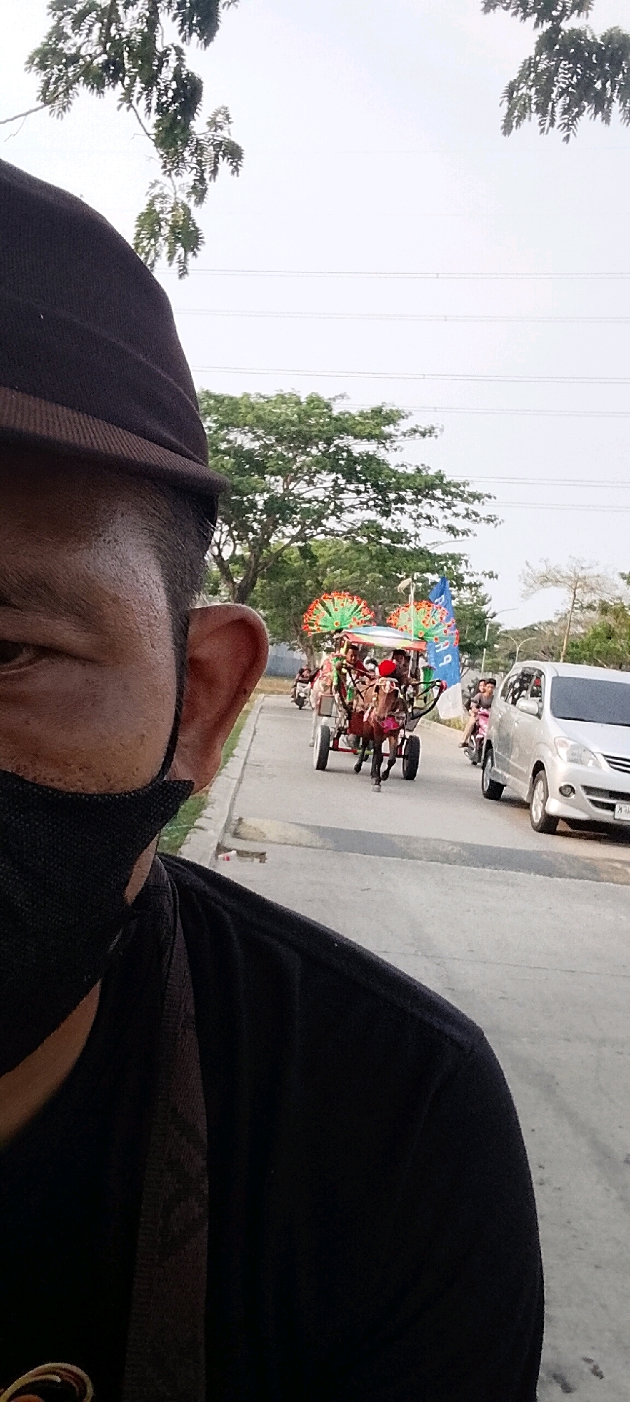 A close-up, low-angle, outdoor, daylight, street-level shot captures a man from the chest up, facing right, with his left side of face and ear visible. He is wearing a dark hat and a black face mask, with his black t-shirt filling the foreground. In the mid-ground, a horse-drawn carriage with people and decorative fans is traveling on a paved road. A silver car is to the right of the carriage, and several motorbikes are in the background. Trees and power lines are visible in the background. The scene is consistent with a procession or parade taking place in a suburban or semi-urban area.