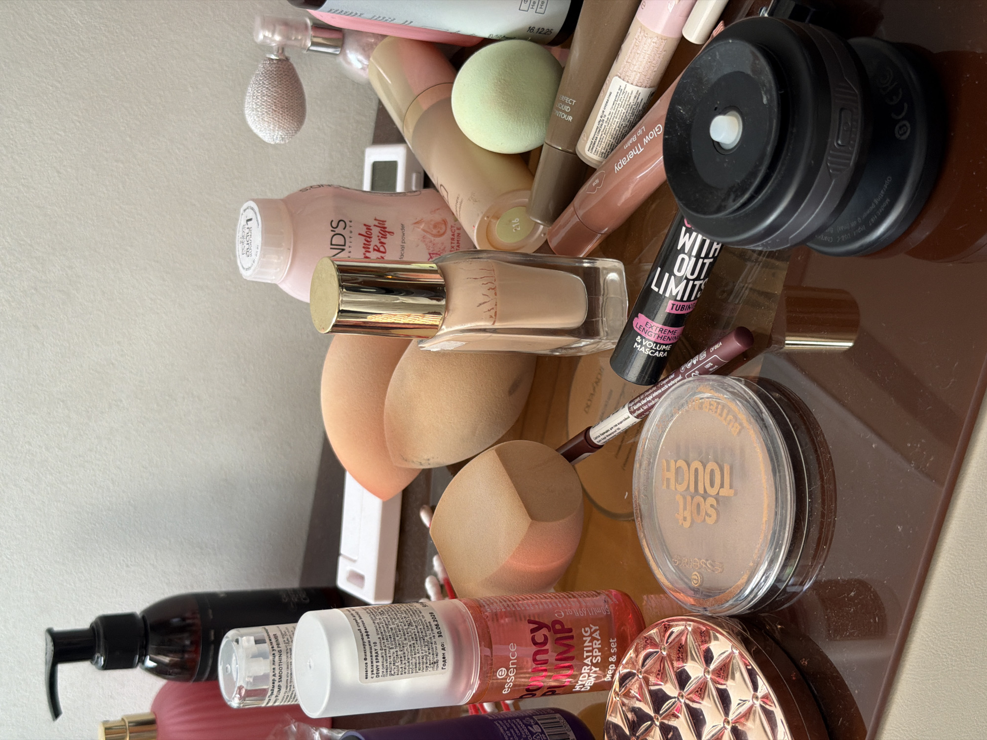 An assortment of makeup products is displayed on a reflective brown surface, likely a vanity or dresser, against a plain white textured wall. The scene appears to be an indoor setting, possibly a residential bathroom or dressing area. The objects are arranged in a somewhat cluttered, yet organized manner, suggesting recent use or preparation for use.
Dominating the foreground is a circular compact powder case with a clear lid, revealing a bronzer labeled "essence SOFT TOUCH." To its left, a rose-gold colored mirrored compact with a geometric design is partially visible. In front of these, a dark brown lipstick or lip liner pencil lies horizontally, with its tip pointing towards a tube of mascara. The mascara tube, black with pink and white lettering, reads "WITH OUT LIMITS EXTREME LENGTH & VOLUME MASCARA."
Slightly behind these items, a glass bottle of foundation with a gold cap stands upright. Next to it, a light pink powder container with a blue lid is visible, labeled "POUDS" and featuring text that appears to be "Melon & Bright." To the right of this, a small, light green cosmetic sponge rests. A small, silver digital device with a white screen is also present, positioned between the powder and a large black pump bottle. This pump bottle, likely a toner or cleanser, is dark brown with a black pump mechanism.
Further back, several makeup sponges in various shades of nude and beige are scattered. A clear spray bottle, labeled "essence DEWY SPRAY" and indicating it is a "HYDRATING DEWY SPRAY prep & set," is prominently placed. Next to it, a pink bottle with a decorative textured surface is partially visible. A clear glass perfume atomizer with a silver sprayer also rests in the background. Other items, including a small, black circular device with a white button and a reflective surface, and several tubes of cosmetic products, are interspersed throughout the arrangement.
The lighting is diffused, suggesting ambient room light or soft artificial lighting, casting subtle shadows on the reflective surface. The overall impression is one of a personal collection of beauty products, neatly arranged for use.
Based on the information from the original image, the setting is Krasnodar, Russia.