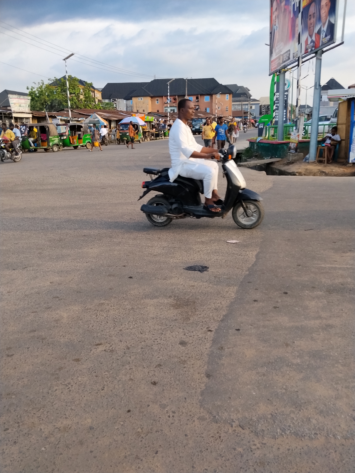 The image depicts a man in white attire riding a black scooter on a paved road in Aba, Nigeria. He is heading toward the right side of the frame. In the background, there are various buildings, commercial stalls, and people. Tricycles (keke) are also visible along the roadside. A large billboard is present on the right. The sky is overcast with clouds. The road surface appears worn.