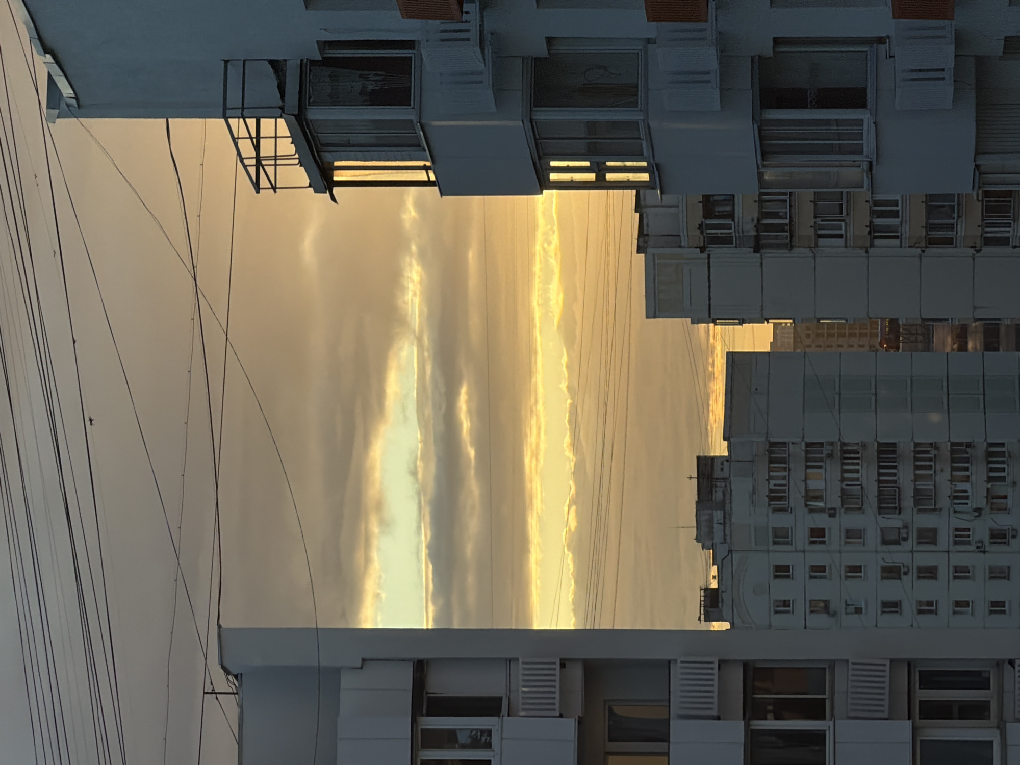 The image shows a view of apartment buildings in the city of Khimki, Russia, at sunset. The sky is filled with dramatic clouds, illuminated by the golden light of the setting sun. Power lines crisscross the sky in the foreground, with the tops of several multi-story apartment buildings visible. The buildings are characterized by their uniform, block-like architecture, typical of Soviet-era housing. Balconies and windows are visible on the facades of the buildings. The overall impression is one of a mundane urban landscape softened by the beauty of the sunset. There are no people visible in the image.