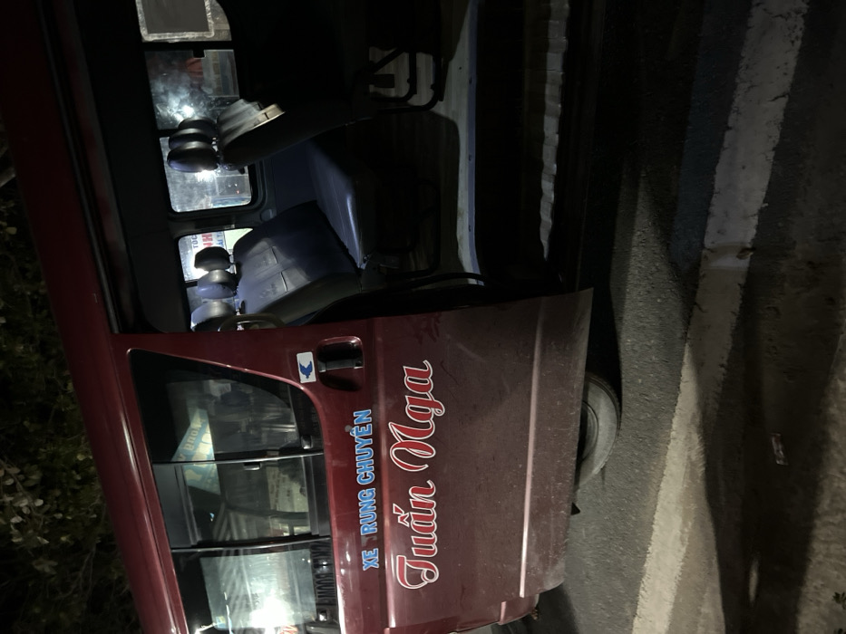 A maroon-colored vehicle, likely a van or small truck, is parked by the side of a road at night. The image is captured from a side angle, showing the vehicle's driver's side door and front window, with part of the road visible to its right. The setting is dark, suggesting nighttime, with reflections of interior lights or distant light sources visible through the windows.

Inside the vehicle, through the glass, parts of the dashboard, seats, and some reflective metallic objects are dimly visible. The exterior of the vehicle appears somewhat dusty. On the maroon side door, there is text written in Vietnamese: "XE RUNG CHUYỂN" in blue lettering and below it, "Tuấn Nga" in a more ornate red script with a white outline. A small blue and white sticker featuring a chicken icon is also present on the door, near the handle.

To the right of the vehicle, the dark asphalt road is visible, marked with two parallel white lines, one thicker than the other, running vertically in the frame (horizontally on the road surface). The vehicle's front tire is partially visible near the bottom right of the door. The overall scene suggests the vehicle is stationary on a paved surface, likely on a street or roadside in Hà Tiên, Vietnam, given the provided location context.