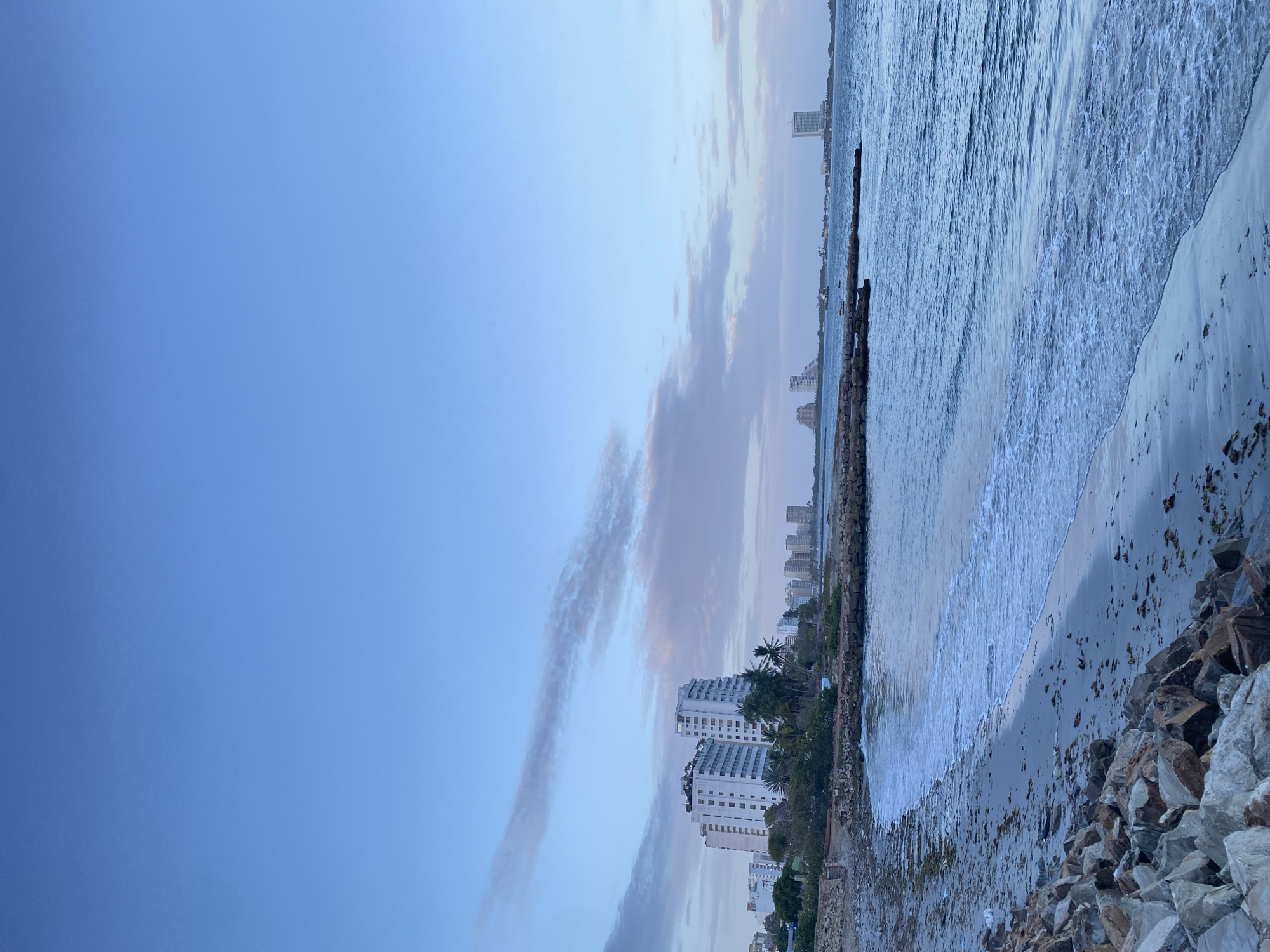 The image depicts a coastal scene in Porlamar, Venezuela. The sky is predominantly blue with streaky clouds. The shoreline consists of a beach with waves breaking onto the sand, alongside a rocky embankment. In the background are several high-rise buildings, indicative of urban development. The perspective suggests the photograph was taken at ground level, close to the rocky shore.