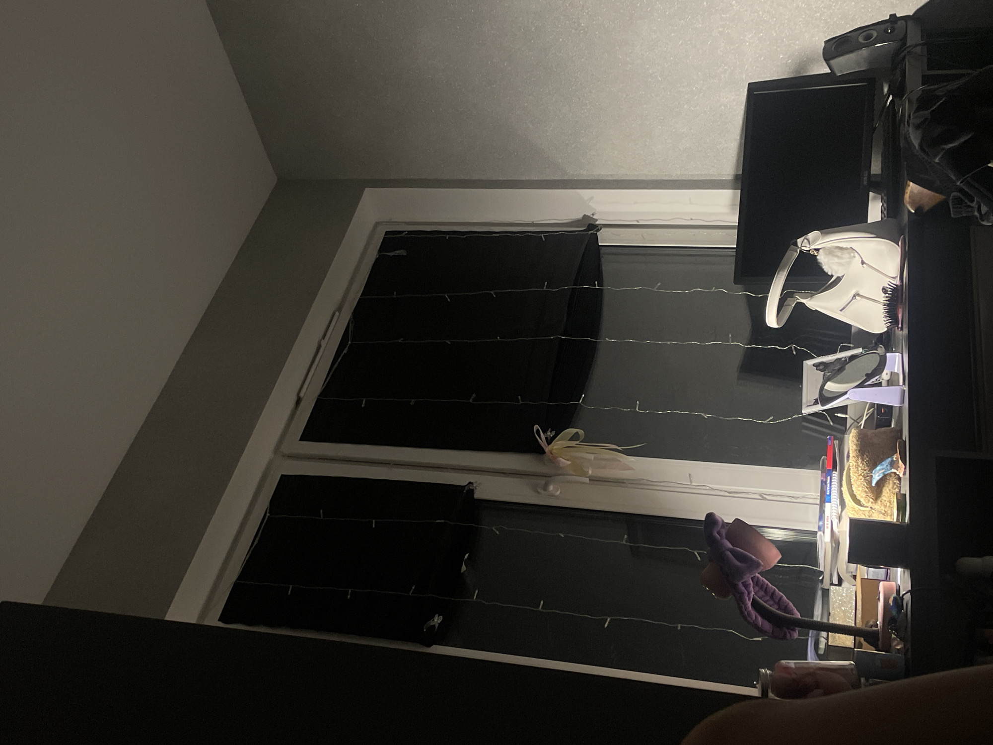 The image shows a dark indoor scene with a window and a desk. The window has two black curtains partially drawn and adorned with fairy lights. A white purse is on the desk, along with a monitor and speakers. Other items on the desk are various toiletries, including makeup brushes. The wall and ceiling are painted in shades of gray. No people are clearly visible in the image, though a body part is shown. The location is likely Saint Petersburg, Russia, but that cannot be determined from the image itself.
