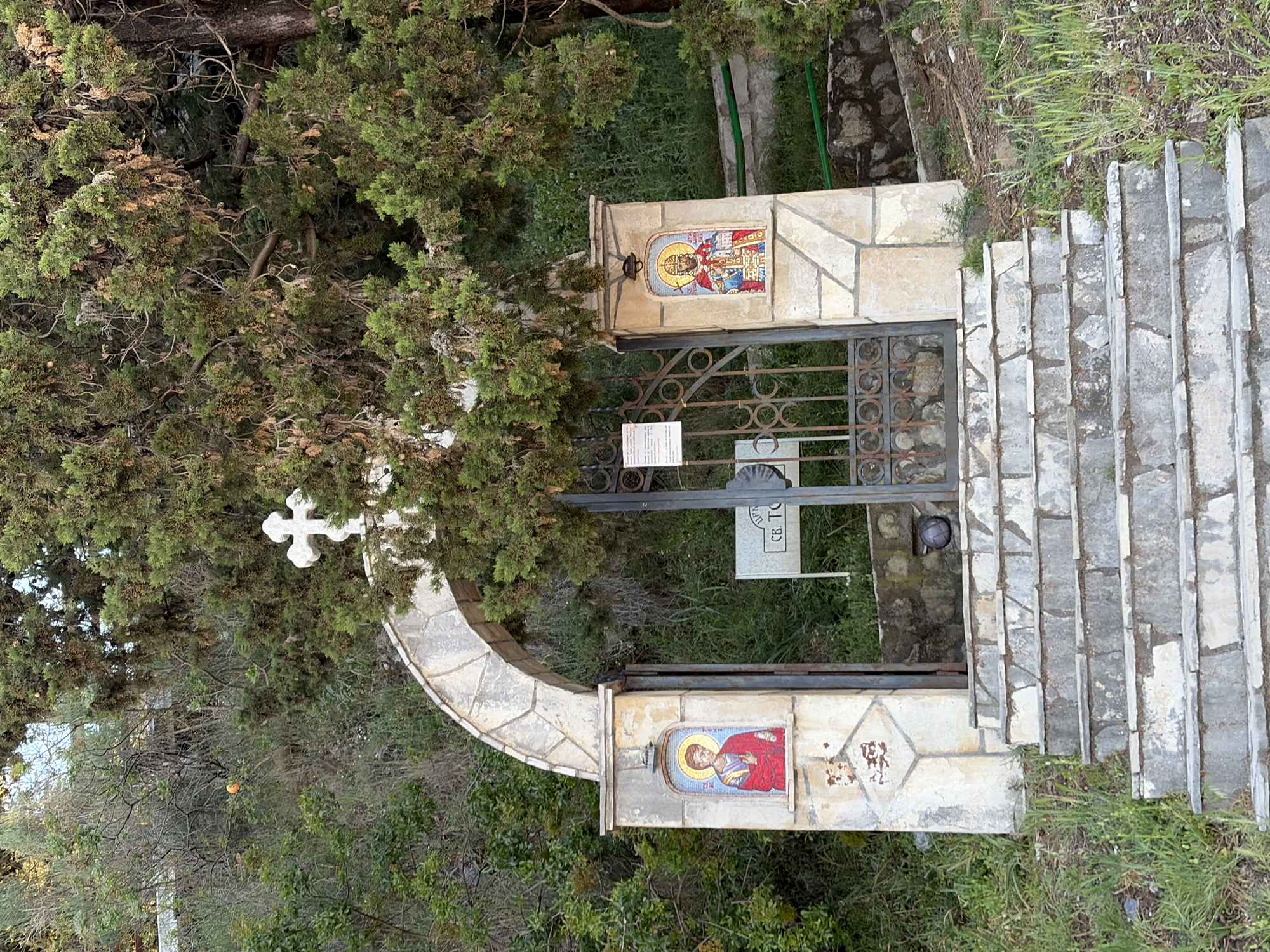 A ground-level, vertical-format shot presents a stone and wrought-iron entrance gate in Boreti, Montenegro, flanked by lush greenery.
A stone archway supports a prominent white cross on the left side of the gate. The gate itself is constructed of wrought-iron, decorated with circular and semi-circular motifs. Stone pillars on either side of the gate display religious mosaics depicting figures with halos. One mosaic shows a figure clad in red robes, while the other portrays a figure in multi-colored garments.
The entrance is bordered by a long flight of stone steps leading uphill on the right side. Grass and vegetation grow along the edges of the stairs and around the gate. Behind the gate, more greenery is visible.