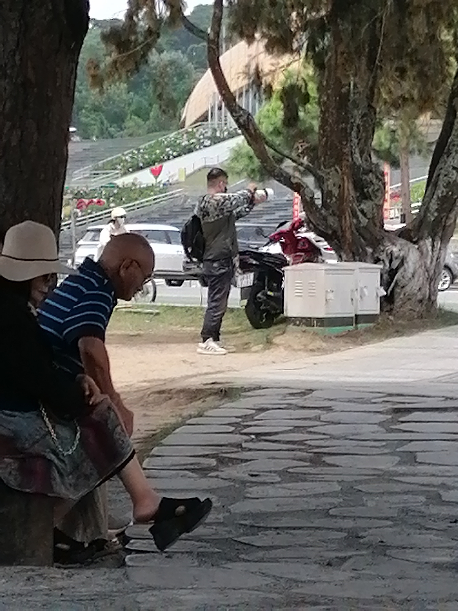 In Da Lat, Vietnam, at the Xuan Huong Lake area, an elderly Vietnamese man with glasses and a shaved head, wearing a blue and white striped shirt, is seated on a bench. Beside him, partially obscured, is an individual wearing a wide-brimmed hat and dark clothing. In the background, a man in a camouflage jacket and dark pants stands near a motorcycle and a white electrical box. A white car is also visible behind him. The environment features large trees and a paved walkway composed of irregular stone slabs, suggestive of a park or public area.