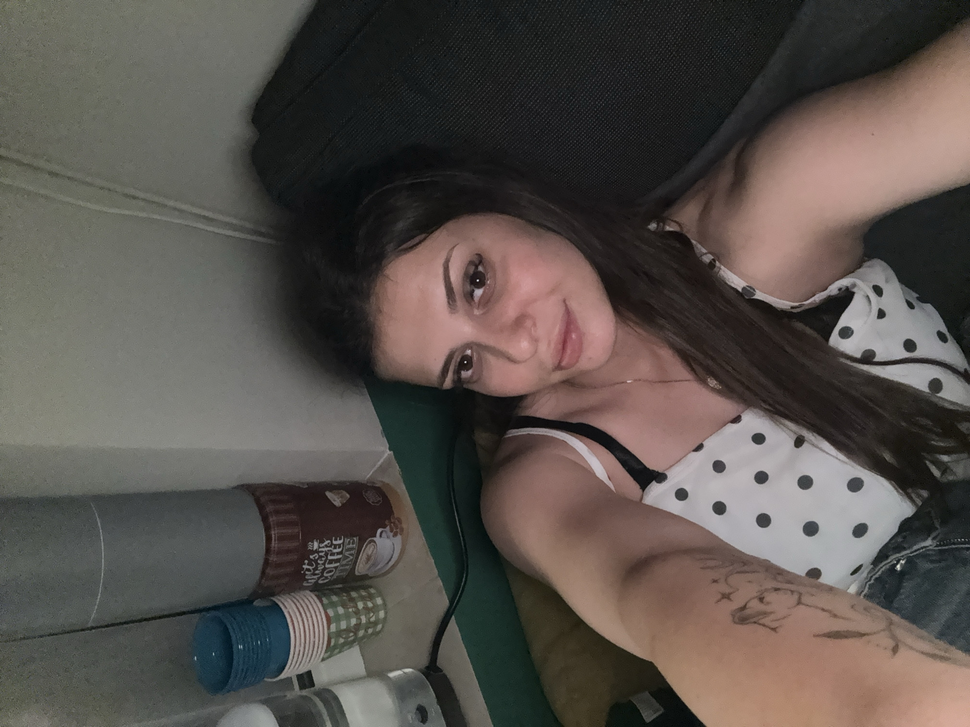 A young woman with long, dark hair is lying down, looking at the camera. She is wearing a white top with black polka dots and a black bra. She has a tattoo on her left arm. In the background, there are stacks of colorful cups and a canister that reads "It's always coffee time." The lighting is dim, suggesting an indoor setting, possibly a home. There are no identifiable landmarks or clues that would place the image in Plovdiv, Bulgaria.
