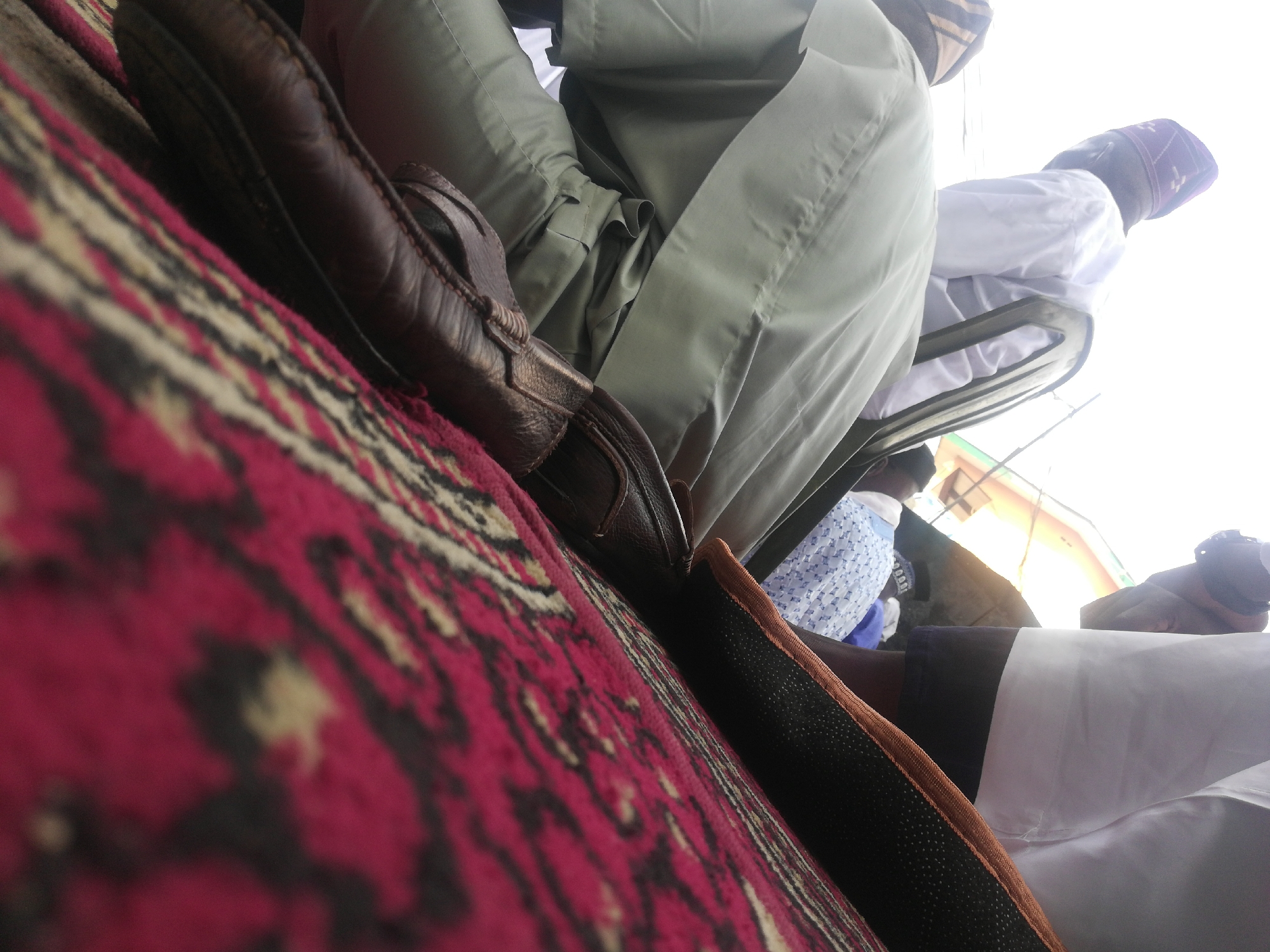 The image depicts a seated individual wearing an olive-green robe or garment, with their legs crossed. A brown loafer shoe is visible on the edge of a vibrant, red, black, and beige patterned carpet. In the background, another person is partially visible, wearing a white shirt and a patterned cap. Additional individuals can be seen in the far background. The environment appears to be an outdoor setting, possibly within an urban or communal area.