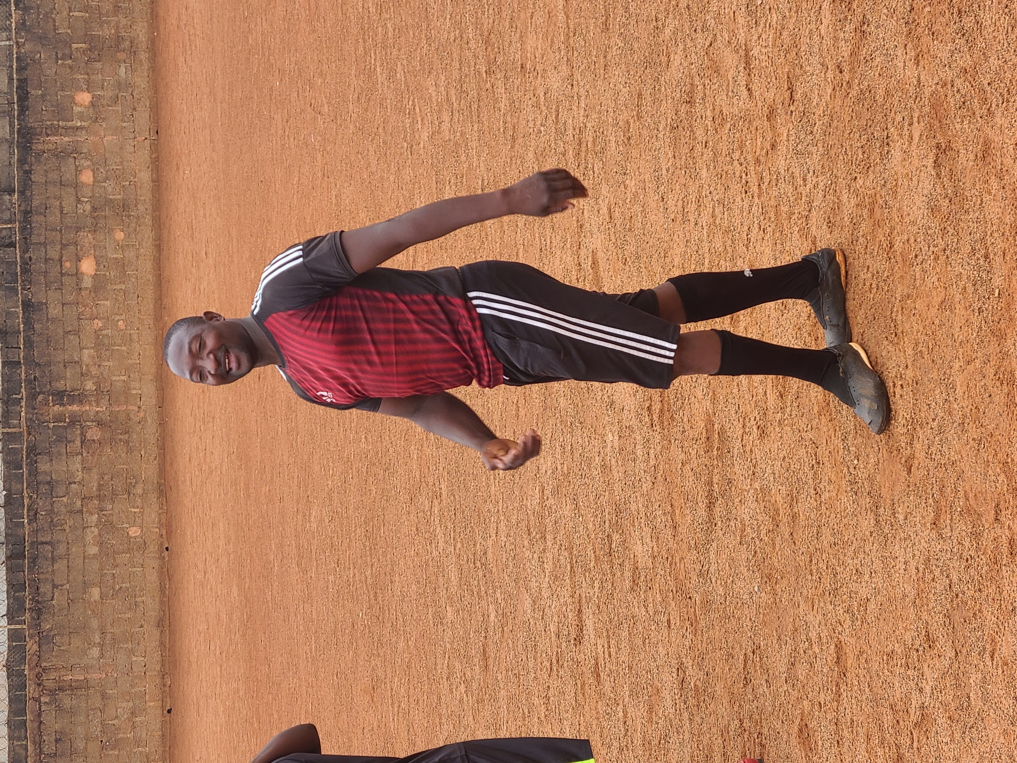 A man stands on an outdoor ground, wearing a red and black athletic jersey with three white stripes on the black shorts. He has on black socks and dark athletic shoes. The ground is textured and reddish-brown. A brick wall is partially visible to the left.