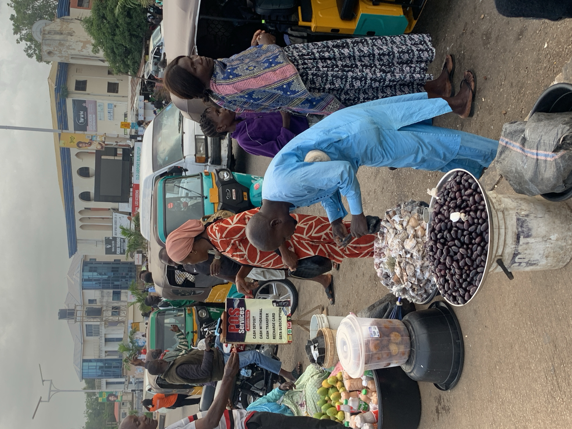 In Barakin Lerawi, Nigeria, a busy street scene unfolds with multiple individuals and vehicles. In the foreground, a person in a blue robe is bending over a large white bucket filled with dark, oval-shaped fruit, possibly olives, which are arranged on a metal plate. To the left, several plastic containers and buckets hold produce, including green fruits that appear to be mangoes, and smaller brown items. A signpost with the text "POS SERVICES" and a list of services such as "CASH DEPOSIT," "CASH WITHDRAWAL," and "DATA SUBSCRIPTION" stands nearby, indicating a service point. Several auto-rickshaws, a common mode of transport in the region, are present. One green and yellow auto-rickshaw is in the center, with a person in a patterned red and white dress standing next to it, looking towards the "POS SERVICES" sign. Further back, a white van and other vehicles are visible. In the background, buildings with modern architectural features, including glass windows, are seen, suggesting a commercial area. The sky is overcast, and the ground is paved with dirt and some debris. The atmosphere suggests a marketplace or a busy intersection where commerce and transportation converge.