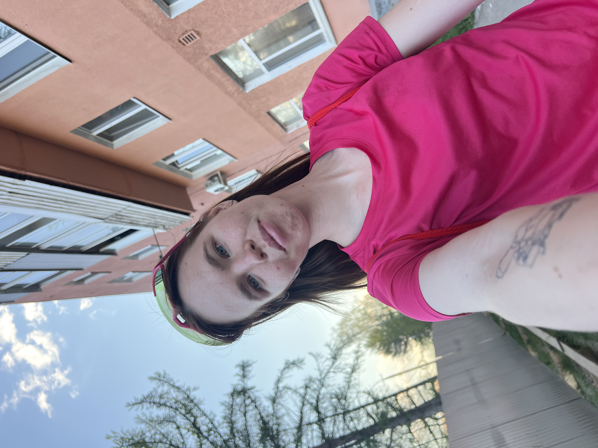 A selfie was taken on a summer day in Perm, Russia. The subject, a person with fair skin, blue eyes, and brown hair, wears a pink t-shirt and a colorful headband. They are standing outside, near a building with multiple windows and a pastel-colored facade. In the background are trees and a partially visible sky. The subject has a tattoo on their arm and is likely holding the camera.