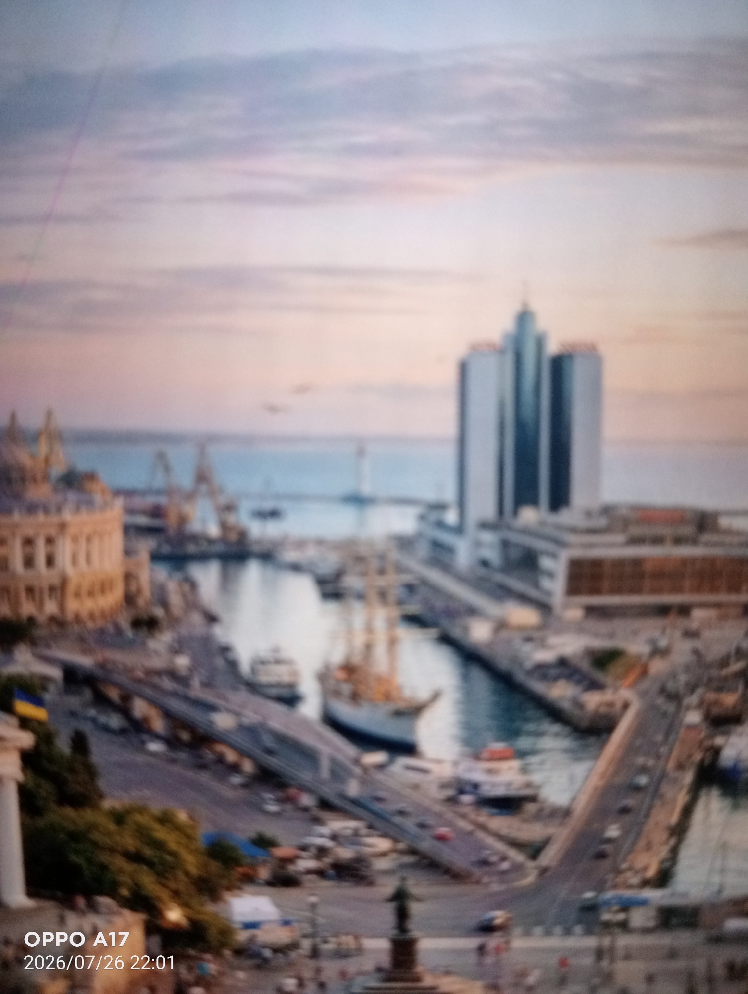 The image is a blurry, telephoto shot of a city harbor at sunset. In the foreground, a Ukrainian flag is visible on a building. A large, modern skyscraper dominates the background, with several boats and cranes in the middle ground, suggesting a busy port. The sky is filled with soft, pastel-colored clouds. The timestamp "2026/07/26 22:01" is superimposed on the lower left corner. The location is identified as Құмөзек, Kazakhstan, however, the architecture and the presence of the Ukrainian flag do not align with this identification. The scene depicts a coastal urban area with significant maritime activity.