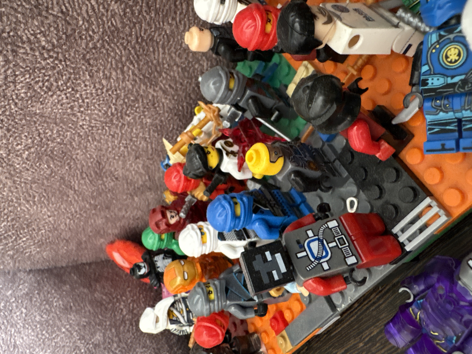 The image shows a collection of Lego minifigures arranged on a textured brown surface, resembling a wall or backdrop. The minifigures are varied in design, with some appearing to be characters from the Lego Ninjago franchise. Several minifigures are in red, blue, white, and black, with some wearing helmets and others having visible faces. Some figures hold accessories like swords or staffs. The minifigures are assembled on a Lego baseplate, which is made of interlocking plastic bricks in various colors, including orange, gray, and green. A purple translucent Lego object is visible in the lower left corner. The overall scene is a close-up shot, with a shallow depth of field that blurs the background and emphasizes the Lego figures in the foreground. The lighting is bright, casting subtle shadows.