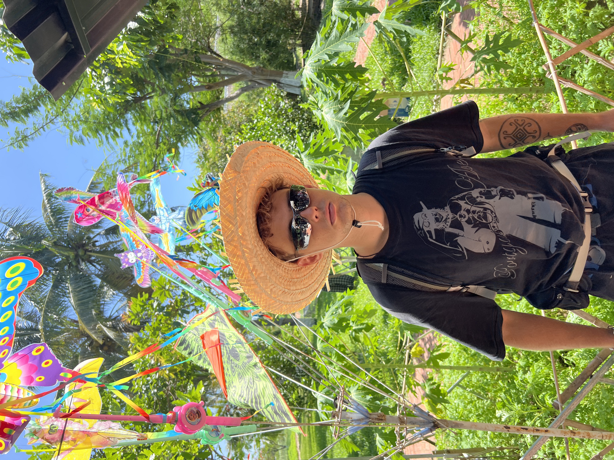 This image captures a young male individual standing outdoors in Nam Cam Ranh, Vietnam. The subject is centrally positioned, facing the camera directly, wearing a light brown straw hat with a chin strap and dark mirrored sunglasses. He is attired in a dark, short-sleeved t-shirt featuring a light-colored graphic design of a figure and stylized text. A backpack with light-colored straps is worn over his shoulders, and a black fanny pack or utility belt with a lighter strap is fastened around his waist. A circular geometric tattoo is visible on his left forearm.
Behind and around the individual is a dense arrangement of colorful kites attached to poles, displaying various forms including butterflies, owls, and abstract shapes, rendered in bright pink, yellow, blue, green, and purple. Multicolored ribbons and streamers hang from these kite structures. A pink bell-shaped object featuring a cartoon character is affixed to a pole on the left.
The environment is characterized by lush green vegetation. Large-leafed plants, consistent with papaya trees, are visible to the subject's right. Palm trees are present in the upper left background, and other trees and dense foliage are scattered throughout the scene. A clear blue sky is visible above the tree line. A rustic wooden or bamboo fence-like structure is in the lower foreground. A section of dark, corrugated metallic roofing material is partially visible in the upper right corner, suggesting an overhead structure. The scene is brightly lit by natural sunlight.