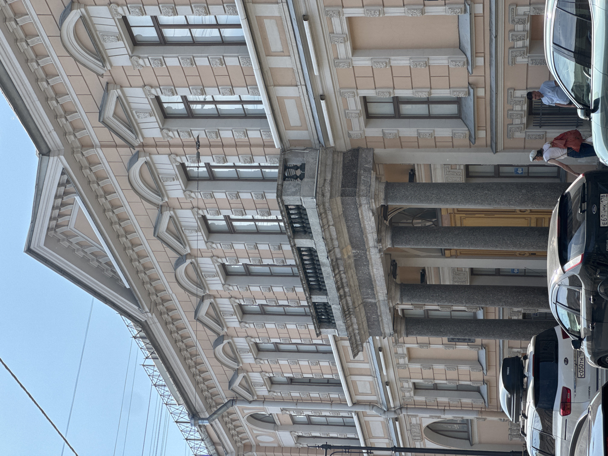 A pale peach building with ornate architectural details is viewed from a street level perspective. The building features multiple stories with evenly spaced windows, many of which are arched or pedimented. A prominent balcony with a stone balustrade and decorative pillars extends across the second story. In front of the building, several cars are parked, including a white SUV with a roof-top carrier and a dark sedan. Two individuals are visible on the sidewalk, one walking away from the camera with a red backpack, and another standing near an entrance. The sky above is clear and blue, with some utility wires visible.