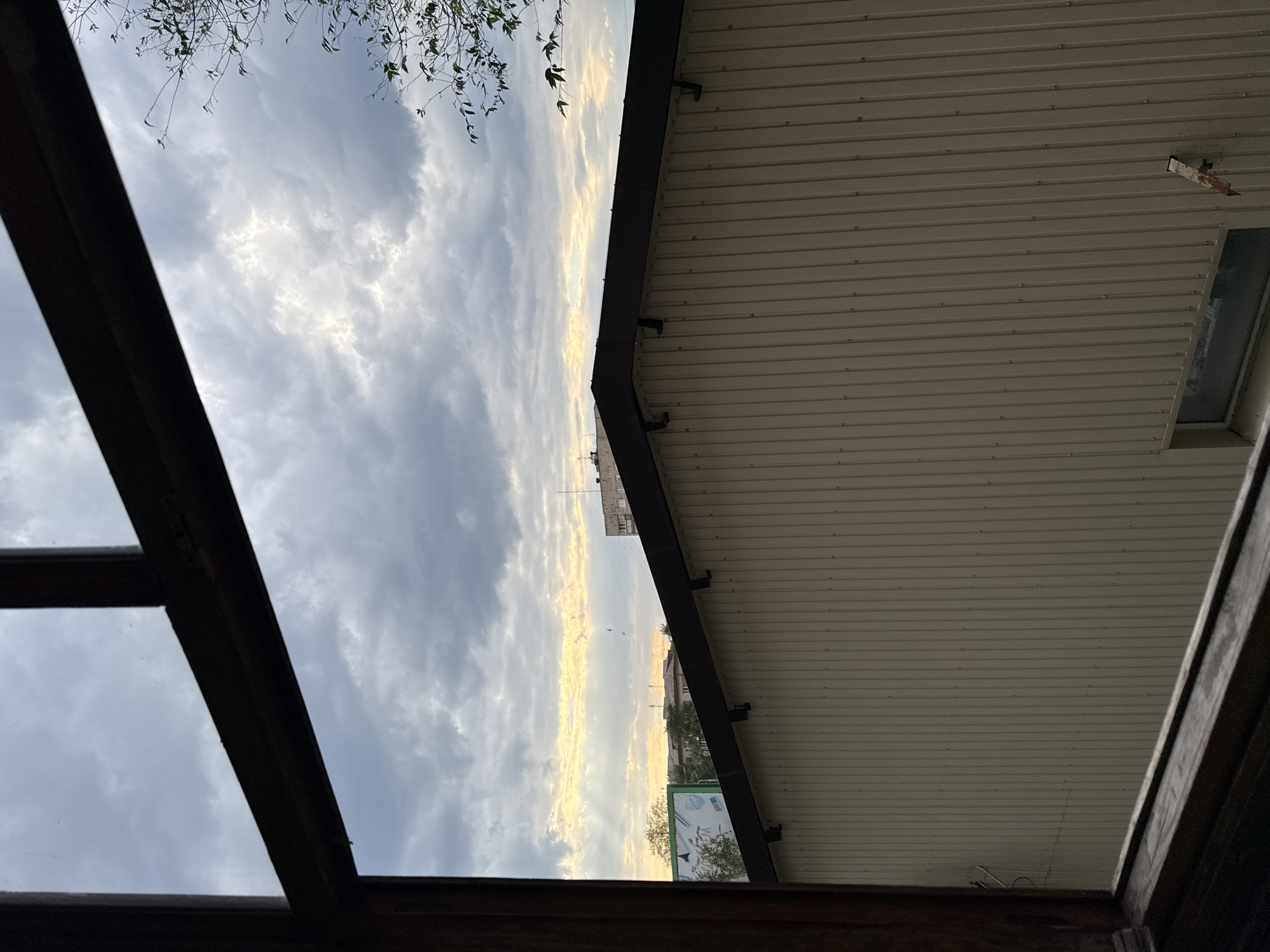 The image presents a view through a wooden window frame, looking outwards at a partly cloudy sky and urban rooftops. The sky exhibits dramatic clouds with streaks of sunlight breaking through, creating a dramatic contrast between light and shadow. Below the sky, a sloped, dark-roofed structure with light-colored, ribbed siding dominates the mid-ground. In the distance, beyond this structure, are glimpses of other buildings and a billboard. The scene appears to be set in Stukalova Balka, Ukraine. There are no people or animals visible in the image. The overall impression is of a somewhat overcast day in an urban or semi-urban environment.