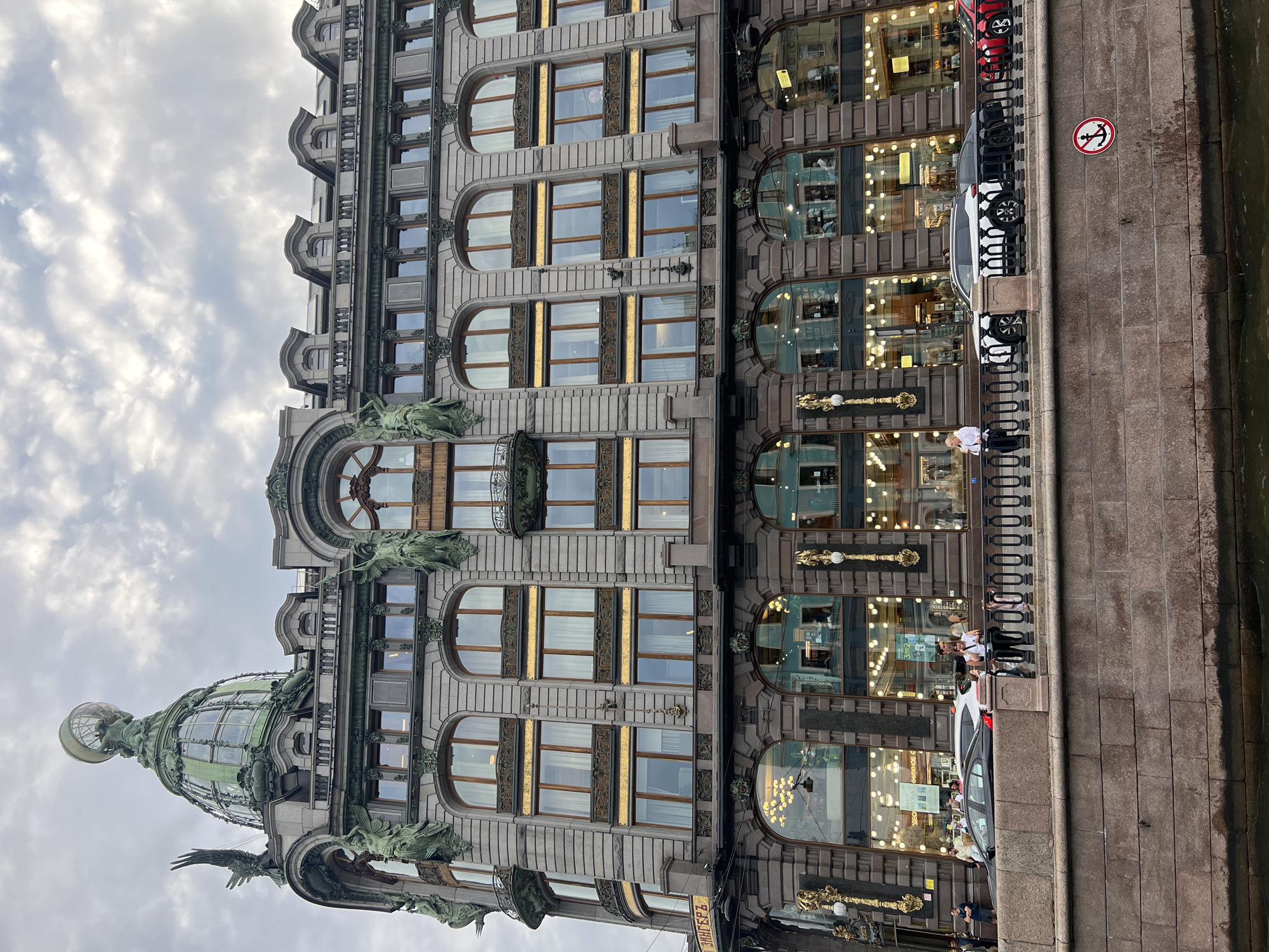 The image depicts a grand, ornate building with a domed roof, situated along a canal in Saint Petersburg, Russia. The building's facade is made of stone and features numerous large windows, many of which are illuminated, suggesting it is either late afternoon or early evening. Architectural details include decorative sculptures and elaborate window frames.
Several people are visible in the foreground, standing along the embankment and near the building's entrance. Cars are parked along the street beside the building, and the canal's water is visible. A circular sign with an anchor symbol crossed out, indicating no anchoring, is posted on the embankment. The sky is overcast with clouds, suggesting a possibly cool or mild day. The overall scene is one of urban life with significant historical and architectural presence.
Visible text includes Cyrillic script on a banner near the bottom center of the image, though it is partially obscured and difficult to read clearly. The signage on the shop windows is also in Cyrillic.