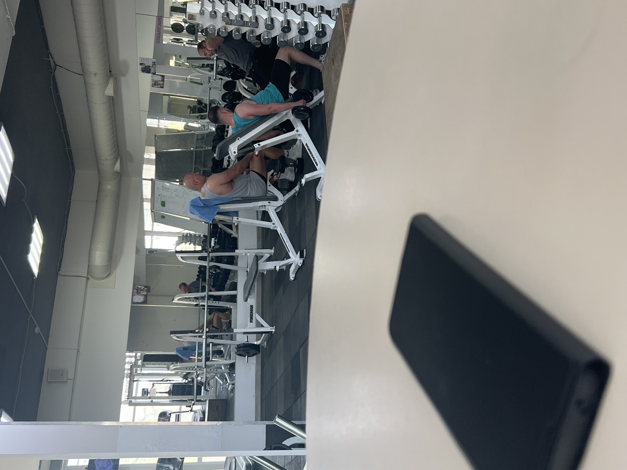 The image captures an indoor gym environment, possibly located in Sochi, Russia, given the context. In the foreground, a dark-colored smartphone lies on a light-colored surface, suggesting it is on a table or counter. The background reveals a gym scene with several individuals engaged in weightlifting exercises. Three men are visible: one in a gray tank top and shorts is seated, another in a teal tank top and black shorts is performing a dumbbell exercise, and a third man in a gray t-shirt and black shorts is also engaged in weightlifting. The gym is equipped with various weight machines, dumbbells, and mirrors, which reflect parts of the gym and its equipment. The lighting appears to be overhead fluorescent fixtures.