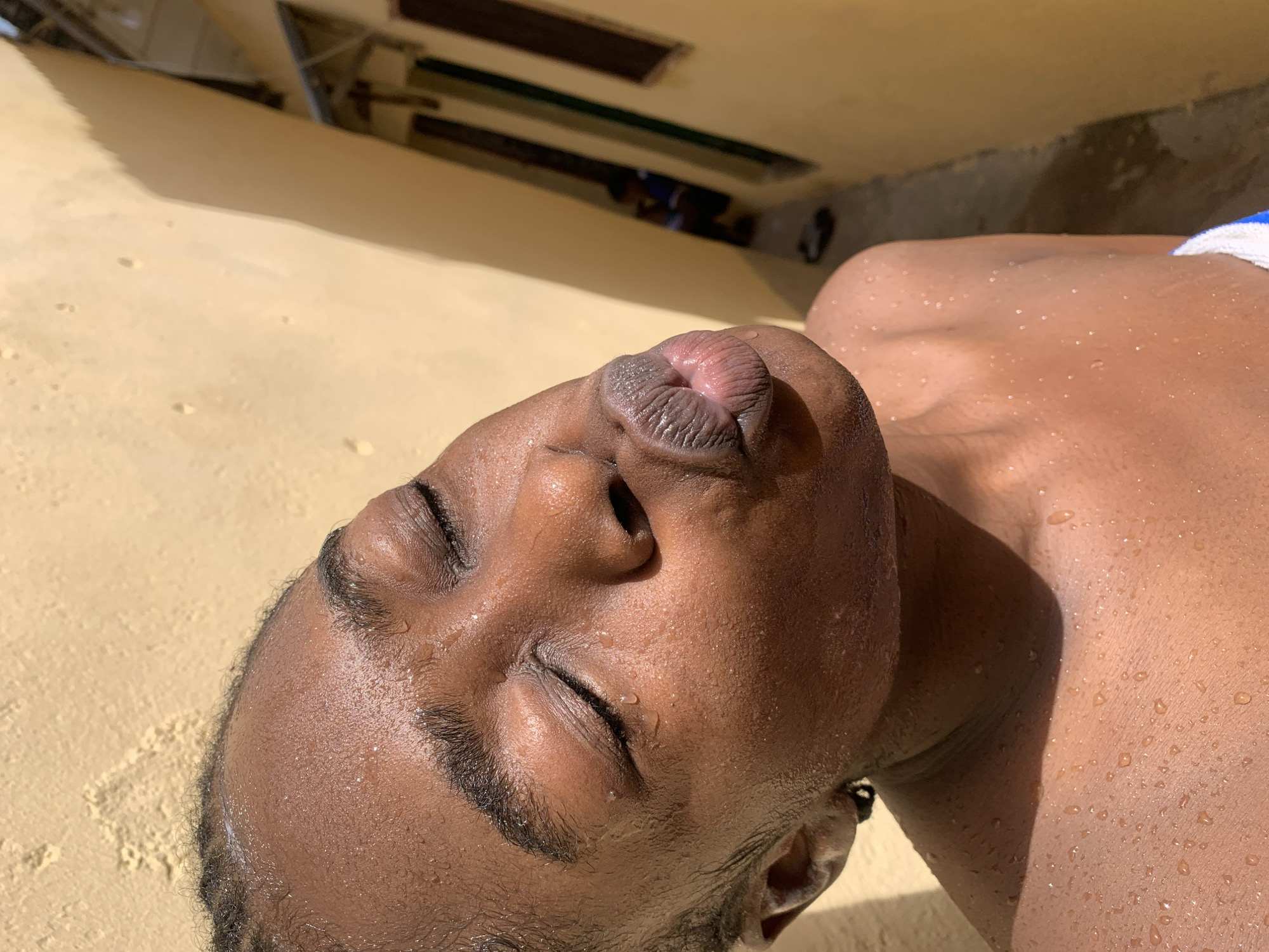 A close-up, eye-level shot shows a young Black individual outdoors, with their eyes closed and lips puckered in a kiss. The subject's face and upper torso are covered in water droplets, suggesting they may have recently been swimming or showered. The background consists of a textured, yellow wall with strong shadows, and a narrow alleyway with buildings on either side. The setting is identifiable as Unguwar Saya, Nigeria, based on prior information. The lighting is harsh and direct, indicating bright sunlight.