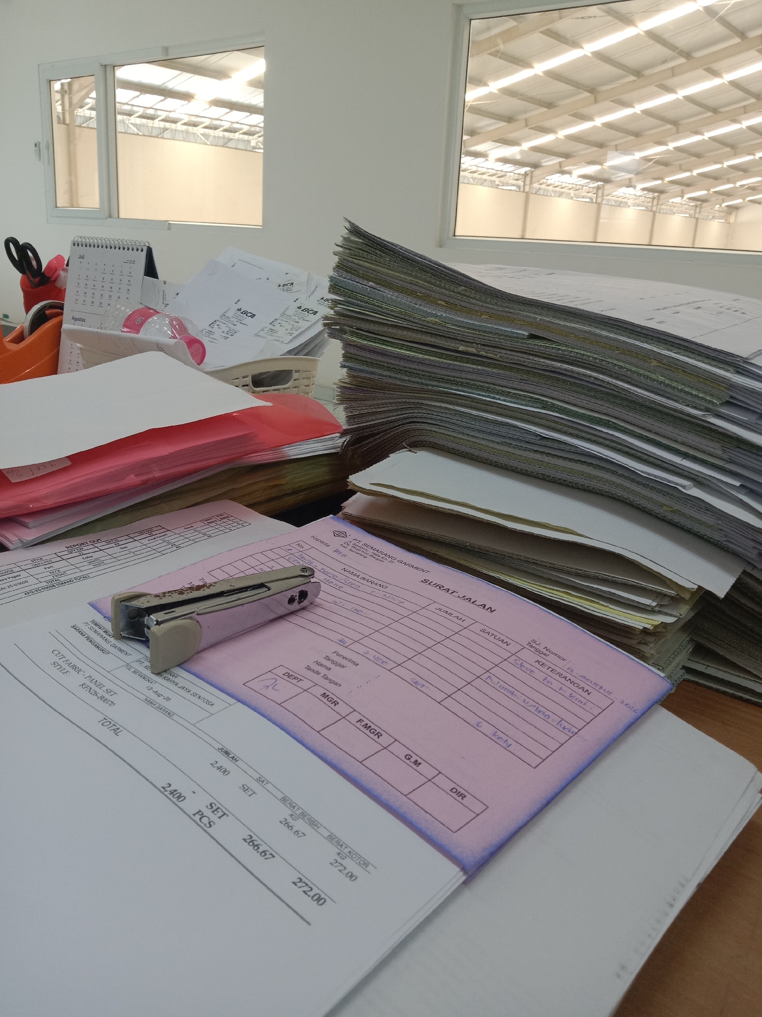 The image captures a desk in a workspace, likely an office, with a large window overlooking an industrial building's interior with a high, well-lit ceiling. The desk is cluttered with stacks of documents, a stapler, a pen holder with scissors and pens, and what appears to be a calendar. One document prominently displayed is a pink form with handwritten entries and printed headings such as "SURAT JALAN" (Delivery Order). This form contains details like "PT. SEMARANG GARMENT" and "KARYA JAYA SENTOSA". Another document shows "CUT FABRIC - PANEL SET" and a style number, "KPN2K-B007D". Below these are columns for "JUMLAH" (Quantity), "SATUAN" (Unit), and "HARGA" (Price), with handwritten entries for "SET" and "PCS". The date "13-Aug-26" and "Agustus" are visible on separate documents, suggesting the setting is Semarang, Indonesia. The overall scene depicts a busy, paper-heavy work environment.