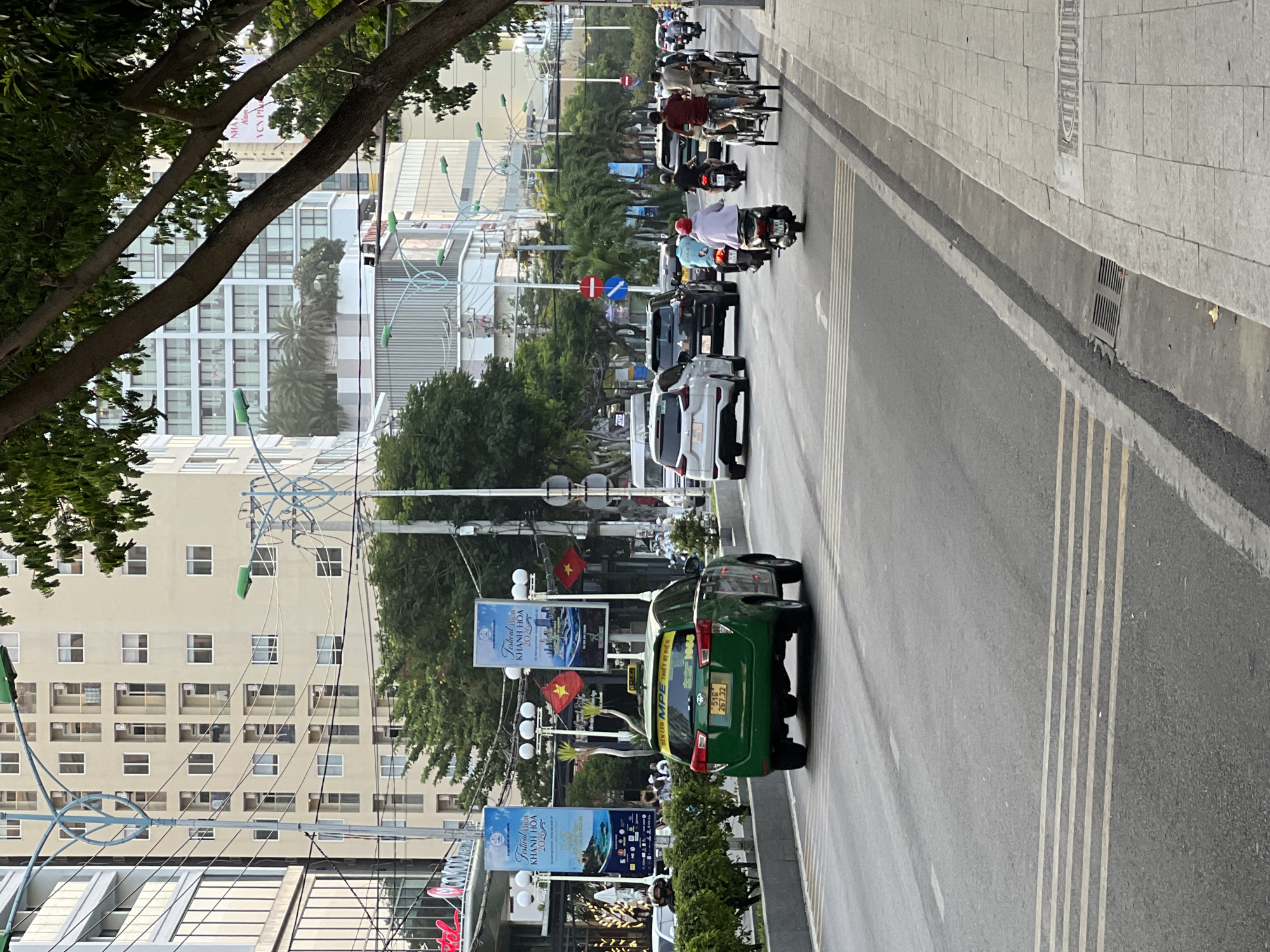This is a street scene in Nha Trang, Vietnam, likely on a busy boulevard, with tall buildings and lush trees lining the road. The primary focus is the traffic on a multi-lane asphalt road. In the foreground, a green taxi with Vietnamese license plates is driving in the left lane. Further down the road, several motorcycles are visible, some carrying single riders and others with passengers. Two men on bicycles are also present, one of them riding a bicycle rickshaw. Cars are also part of the traffic flow. Streetlights and utility poles are scattered throughout the scene, some adorned with decorative elements. Two large banners advertising the "Festival Bien Khanh Hoa 2020" are prominently displayed on poles, each featuring Vietnamese text and images of landscapes. The sidewalks are paved with concrete tiles, and a drain grate is visible near the curb. The overall atmosphere suggests a typical day in a Southeast Asian city, with vibrant urban life and a mix of transportation modes.