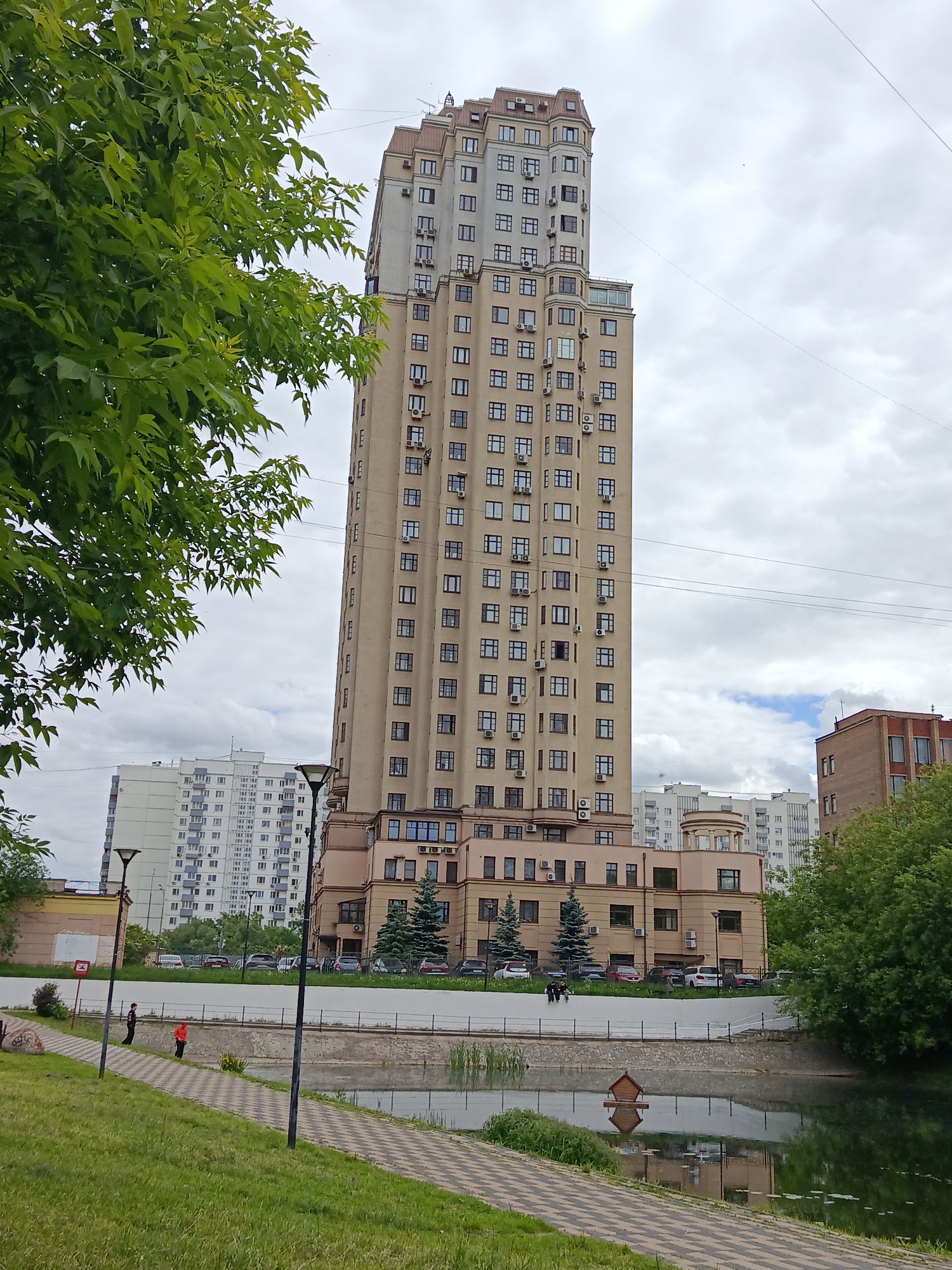The image displays a tall, slender building dominating an urban landscape, with a pond and green spaces in the foreground. The prominent building is light beige/tan, featuring many square windows and numerous external air conditioning units. Its upper sections have varied architectural details and a multi-toned roof.
In front of the building, a calm pond reflects the surrounding greenery and sky. A paved path with a checkered pattern runs alongside the pond, separated from a grassy slope by a low wall and a fence. A small, wooden birdhouse or feeder floats on the water. Two individuals are visible on the path: one in an orange top walking away, and another in a dark top standing further back. A third person is standing near a lamppost further down the path.
The sky is overcast with visible clouds, suggesting daytime. Lush green trees and foliage frame the left side of the image and grow around the pond. In the background, other residential buildings, mostly white or light-colored, are visible, indicating a dense urban setting. A brick-colored building is also present on the far right. Cars are parked in lots behind the main structures. A small red sign with an unidentifiable white symbol is on a post next to the path. The scene depicts a green recreational area within Moscow, Russia.