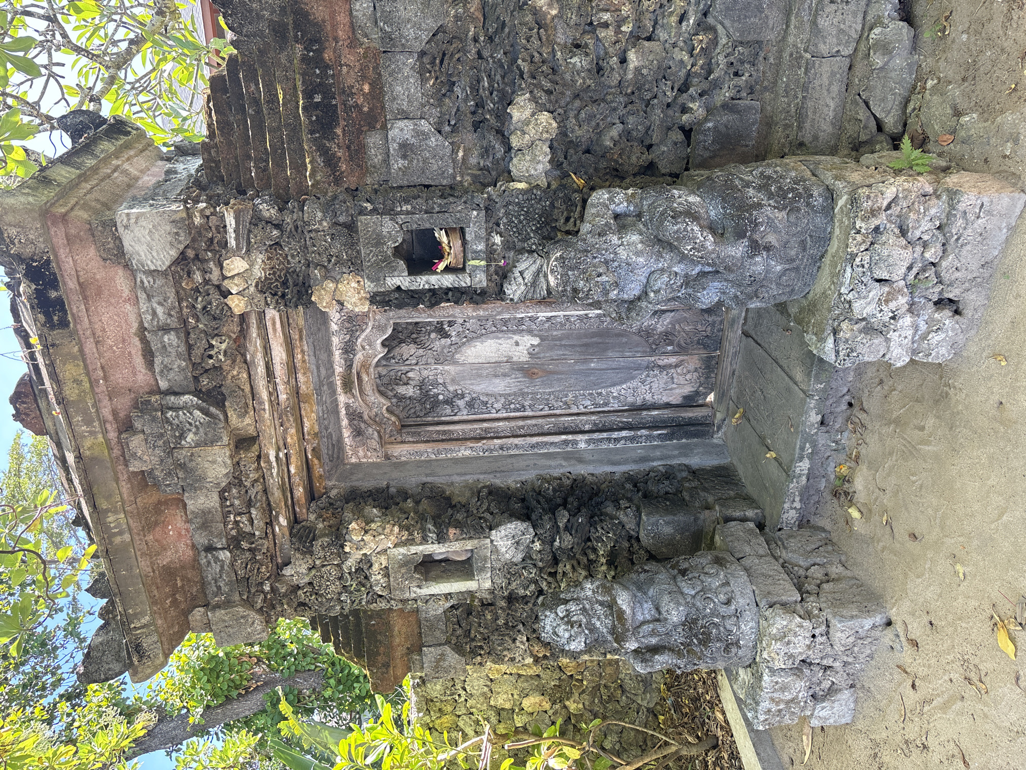The image captures a stone structure in Denpasar, Indonesia, possibly a Balinese temple or shrine. The structure is constructed of rough-hewn gray stone and coral, with carved stone figures flanking a wooden door. The door is closed and weathered. There are small niches in the stonework, one holding what appears to be an offering of flowers and leaves. Green foliage surrounds the base of the structure, contrasting with the gray stone. The ground is covered in light sand or soil.