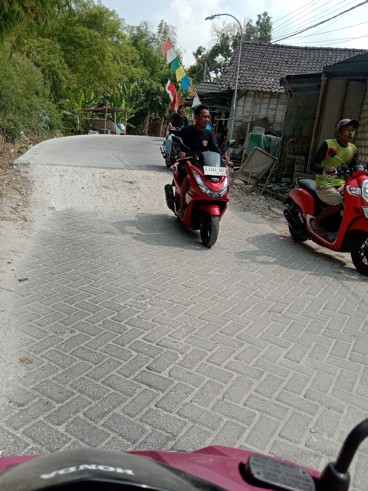 The image depicts a street scene in Jumput, Indonesia, under a clear sky. In the foreground, the handlebars and part of the windshield of a red scooter are visible, suggesting the photo is taken from the perspective of a rider. A red scooter, identified by the license plate "S 4484 ABZ", is the primary subject in the middle ground, ridden by a man wearing a black t-shirt and blue jeans. To the right of this scooter, another red scooter is visible, carrying a person wearing a green shirt and tan shorts. In the background, a cluster of houses with tiled roofs and a street lamp are present. Lush green trees and vegetation line the left side of the road, with several colorful flags strung across a pole. The road is paved with interlocking bricks. The overall impression is one of everyday life in a semi-urban or rural Indonesian setting.