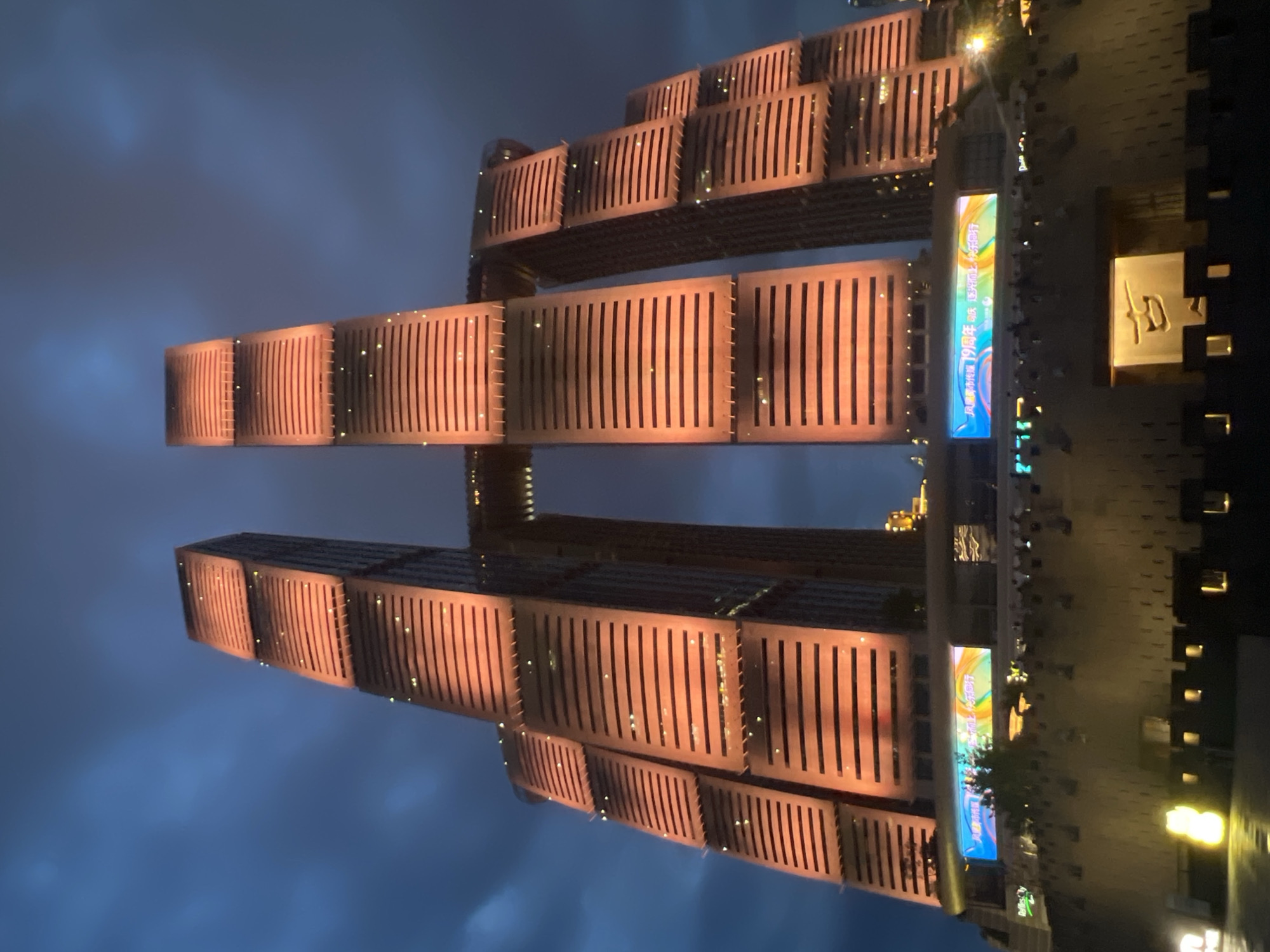 The image shows a close-up, low-angle view of the iconic "Twin Towers" skyscrapers in Yuzhong District, China, at dusk. The buildings are illuminated with warm orange lights, highlighting their layered architectural design and horizontal window bands. A bridge-like structure connects the two main towers at a high elevation. In the foreground, a section of a dark, textured wall with small, illuminated windows forms a lower level. Above this wall, a large digital billboard displays colorful graphics and Chinese text, appearing to celebrate an anniversary. The sky above is a deep blue, with hints of clouds. The overall impression is one of impressive urban architecture and nighttime illumination.