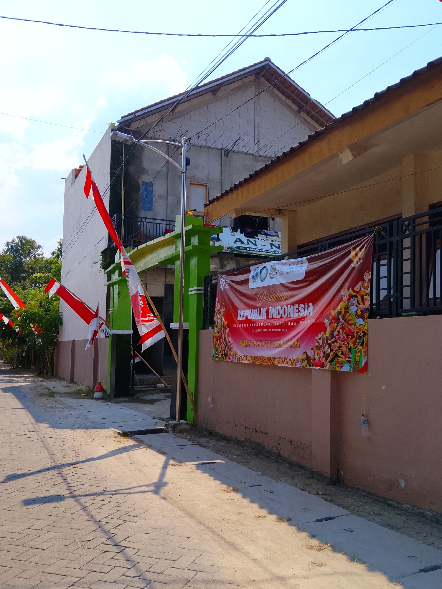 The image depicts a street scene in Padangan, Indonesia, likely near a school, indicated by the banner. The foreground features a paved walkway and a road made of interlocking concrete bricks, casting long shadows from an unseen light source, suggesting it is either early morning or late afternoon. Several red and white Indonesian flags are displayed, some on poles along the walkway and others attached to the building.
The building on the right is a single-story structure with a light brown exterior wall and a yellowish-brown overhang. A dark metal fence runs along the front of the building, behind which windows are visible. A large red banner hangs prominently on the wall, celebrating "Dirgahayu Republik Indonesia" (Independence Day of the Republic of Indonesia). The banner includes text related to the anniversary and "SMKS Pelita Harapan," suggesting it's a vocational school. The banner also features illustrations of people in traditional attire and a Garuda Pancasila emblem.
To the left, a two-story building is partially visible. The lower story has a white exterior wall, and the upper story appears to be constructed from concrete blocks with exposed rebar, suggesting it is either under construction or unfinished. A green gate structure with white trim stands in front of the lower story. An outdoor light fixture is mounted on a pole near this gate. The area is lined with trees and bushes, adding greenery to the scene. In the distance, power lines crisscross the sky.