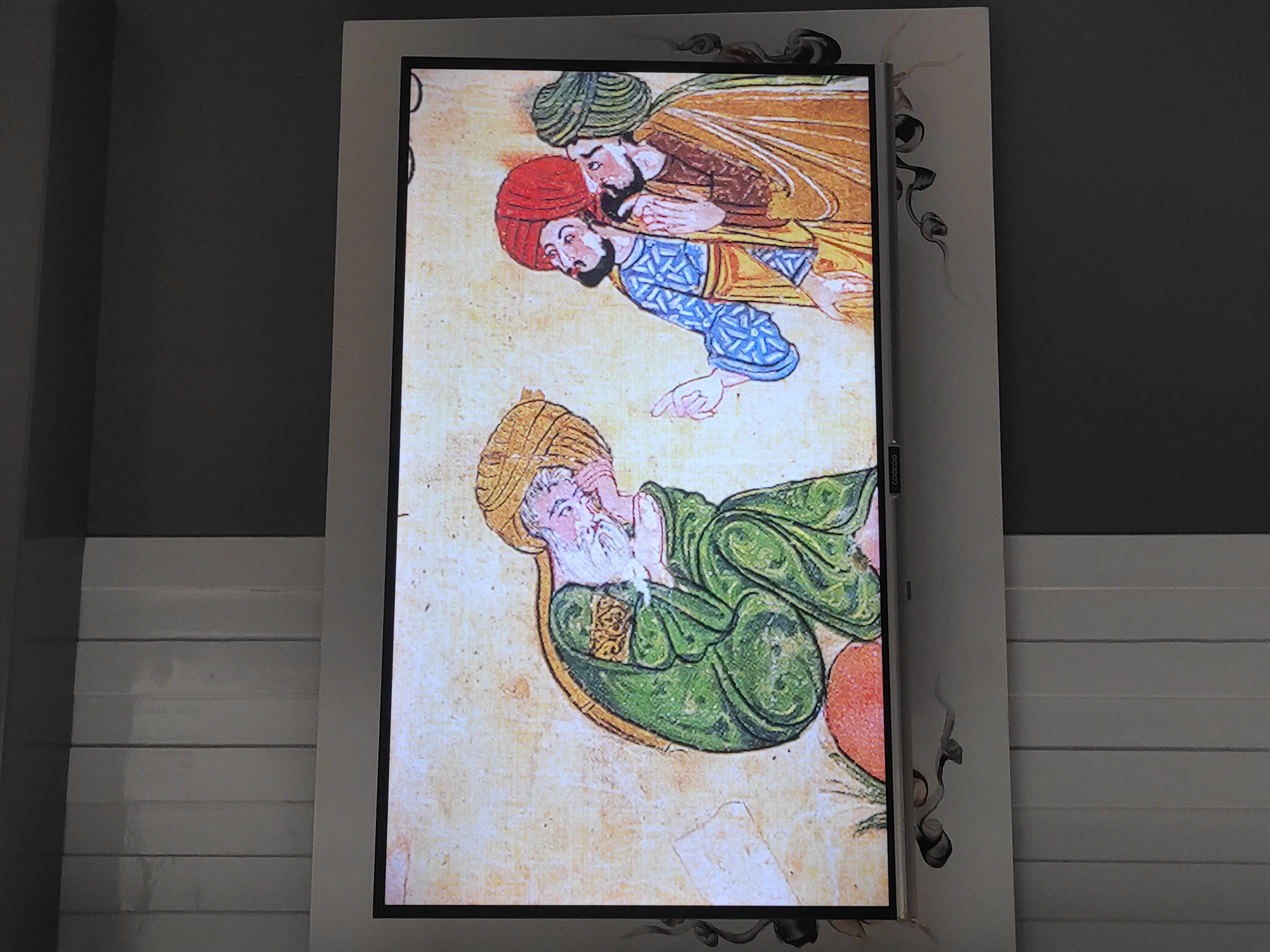 An old, weathered parchment displays an ancient illustration of three men dressed in traditional robes and turbans. On the left, a man with a long white beard and a golden turban is depicted hunched over, with his head resting on his hand. He wears a green robe with intricate gold trim. To the right, two other men stand, both with beards and turbans. One wears a blue robe with a geometric pattern and a red turban, while the other wears a golden robe and a green turban. The illustration is set against a textured, yellowish background with hints of what might be grass or foliage at the bottom. The image is displayed on a television screen with a white frame. The screen is mounted on a wall that appears to be paneled with vertical, glossy white sections on the left and a dark gray section on the right. Decorative dark gray embellishments are visible on the white frame of the television, resembling curling vines or smoke. The overall scene suggests a cultural or historical display, possibly in a public space or a home.