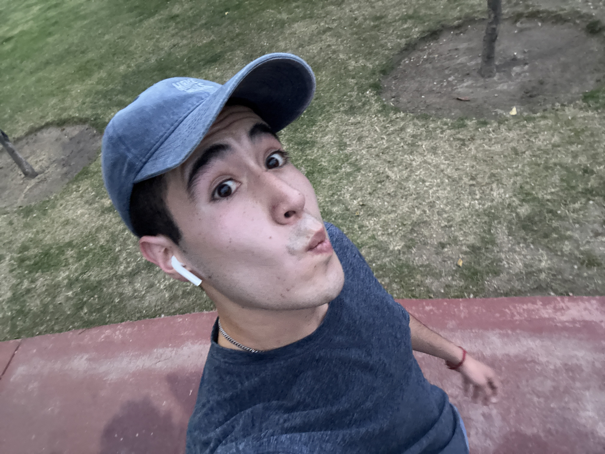 A young man poses for a selfie outdoors, possibly in San Bernardino Tlaxcalancingo, Mexico, although this cannot be verified from the image alone. He is wearing a blue baseball cap, a dark blue t-shirt, and a silver chain necklace. A white wireless earbud is visible in his left ear. He is making a kissing face with wide eyes. He is standing on what appears to be a red-painted concrete surface, with grassy ground and trees in the background. His right arm is slightly extended with a red wristband visible.