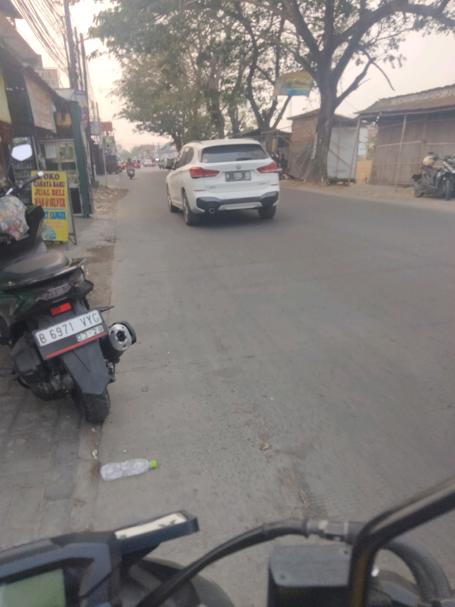 The image captures a street scene in Sepatan, Indonesia, with a focus on a white SUV on the right side of the road and a motorcycle parked on the left. The SUV, a BMW X1, is facing away from the camera and is in motion, indicated by its position on the asphalt road. The motorcycle, a black scooter, is parked on the sidewalk, facing towards the left. Its license plate is clearly visible with the number "B 6971 YYG" and a date of "03-28".
The environment depicts a typical urban setting with shops lining the left side of the street. One shop, with a yellow sign, advertises "Poko Carata Baru Jual Beli Mas & Silver". Trees with lush foliage are present, casting shadows on the road and buildings. Overhead, a complex network of electrical wires crisscrosses the sky, a common sight in Indonesian urban areas. On the sidewalk near the motorcycle, a discarded plastic water bottle with a green cap lies on the ground. The overall lighting suggests it's either early morning or late afternoon, with a soft, diffused light. The road surface is asphalt, showing signs of wear and tear. The background reveals more buildings and vegetation, and other vehicles are visible further down the road, indicating active traffic. The perspective suggests the photo was taken from another motorcycle.