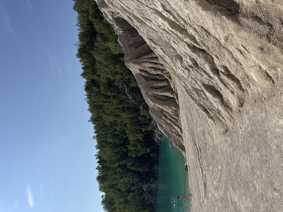 The image displays a sunny, outdoor scene at a large flooded quarry site in Druzhba, Ukraine. Dominating the right and central foreground are vast, light-tan-colored slopes composed of what appears to be sand or clay, marked by distinct, deep erosion gullies and ridges. To the left, a striking turquoise-blue body of water, indicative of a quarry lake, stretches from the foreground to the midground. The far side of the lake is lined by a dense, dark green forest, ascending gently into the background under a clear, bright blue sky with minor high-altitude clouds.
Human presence is observed near and in the water. In the lower-left foreground, a light-skinned individual, appearing female and wearing a light-colored swimsuit, walks horizontally along the sandy shore near the water's edge. Adjacent to this individual, several dark objects, possibly articles of clothing or bags, rest on the sand. Within the turquoise water, at least four other individuals are partially submerged, engaged in swimming or floating activities. The scene is illuminated by strong sunlight, casting defined shadows and highlighting the texture of the eroded slopes.