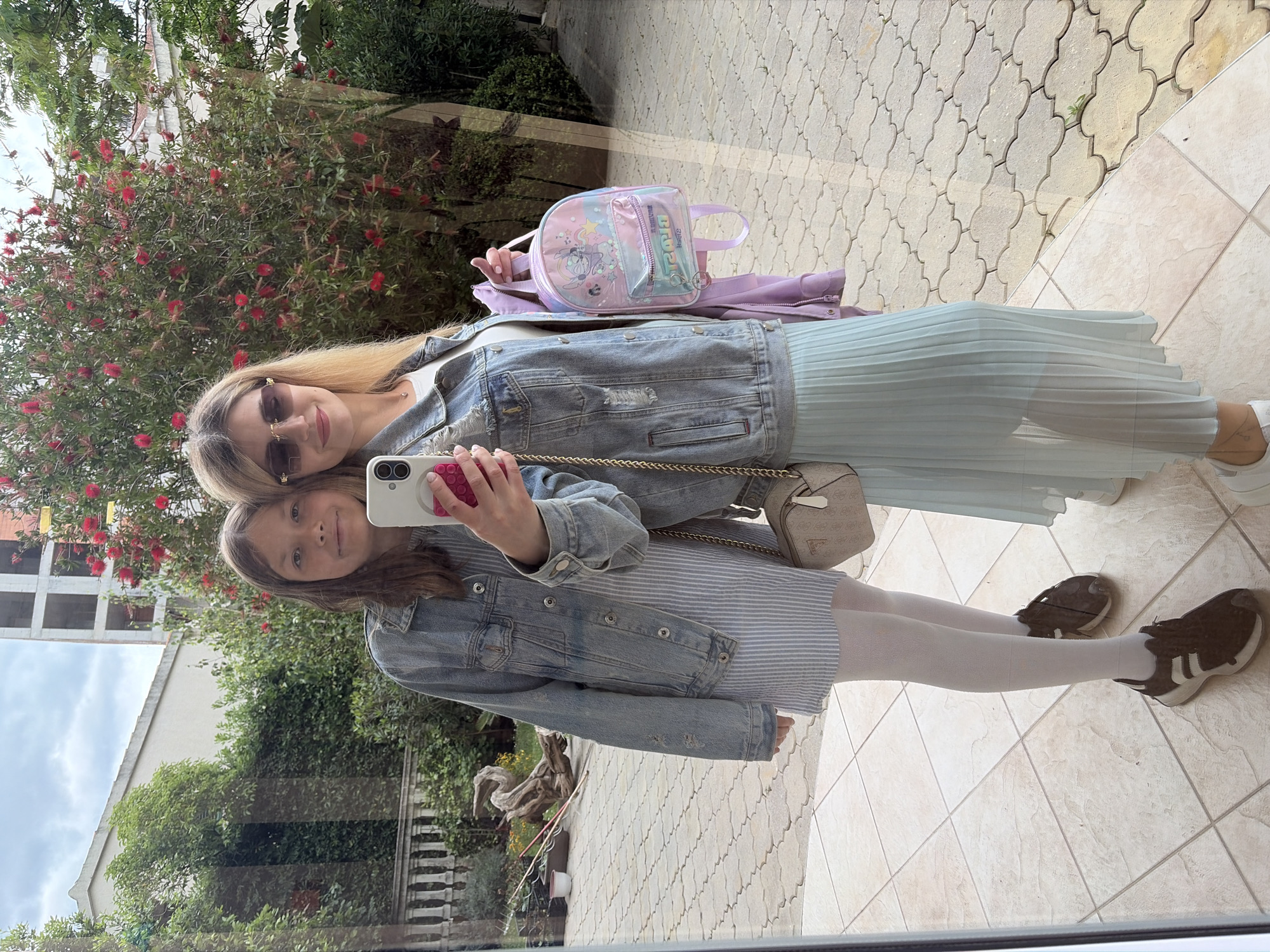 A woman and a girl are posing for a selfie indoors, with a reflection of a street scene visible in the background. The woman, with blonde hair and sunglasses, is wearing a denim jacket, a white top, and a light blue pleated skirt. She has a small handbag with a gold chain strap. The girl, with light brown hair, is wearing a denim jacket over a light-colored striped dress and white tights. She is wearing brown sneakers. A pink backpack is visible next to the woman. The street scene in the reflection includes trees, a building under construction, and the red flowers of a bottlebrush plant. The location is likely in Budva, Montenegro, based on the style of architecture and plants visible in the reflection. The floor they are standing on is tiled.