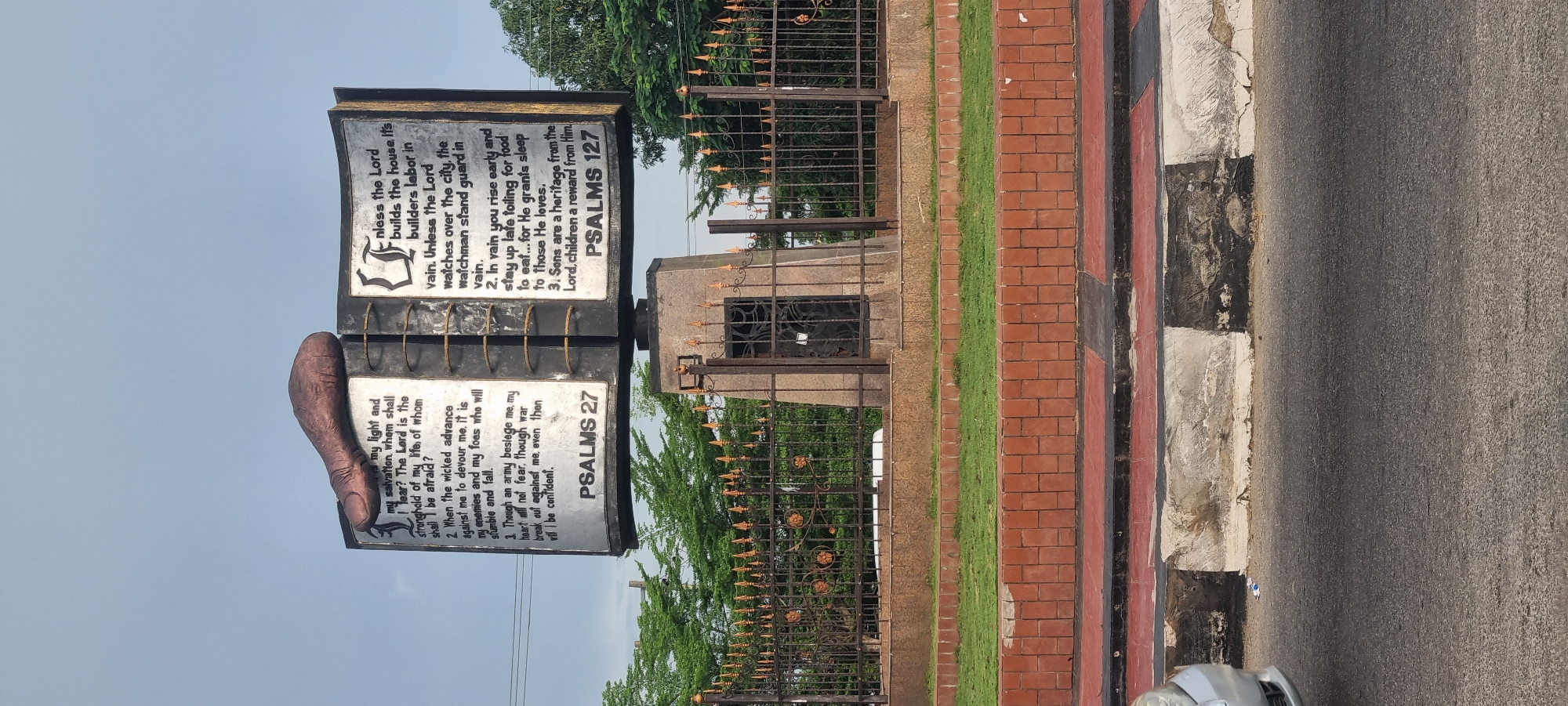 The image depicts a monument in Calabar, Nigeria, displaying excerpts from Psalms 27 and 127. The monument has two vertical stone slabs connected by a metal structure with visible rings. Each slab features engraved biblical verses in English. The first slab displays verses from Psalm 27, while the second slab shows Psalm 127. A hand sculpture is visible on the left slab.
The monument is surrounded by a wrought-iron fence with gold-colored accents and is located near a walkway with alternating sections of red brick and grey concrete. Lush green trees are visible in the background. A portion of the road and a car are visible on the right side of the frame.