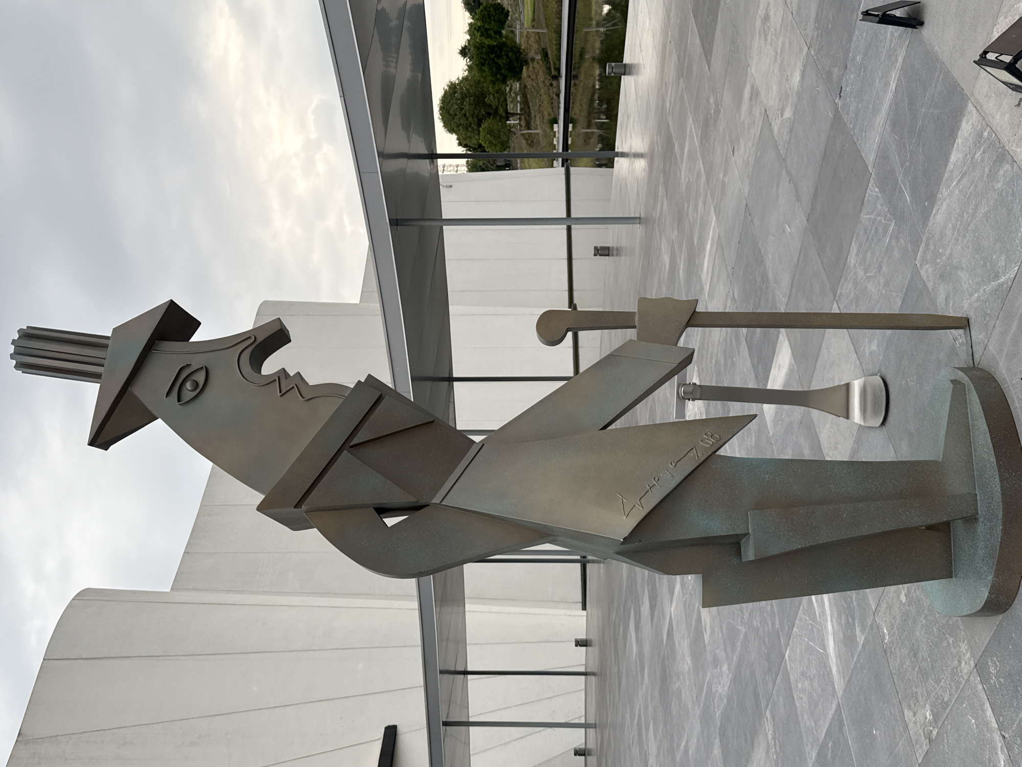 The image shows a bronze statue of a cubist figure standing outside a modern building. The statue has a hat, an eye, and angular features. A sword and another tool rest on its shoulder. It has an inscription that appears to read "L'apres 7.08". The statue stands on a square stone platform, and the ground is covered in a diamond-patterned stone tile. In the background, there is a modern building with white walls and glass railings. Beyond that, there is a grassy area with trees and a body of water. The location is San Bernardino Tlaxcalancingo, Mexico.